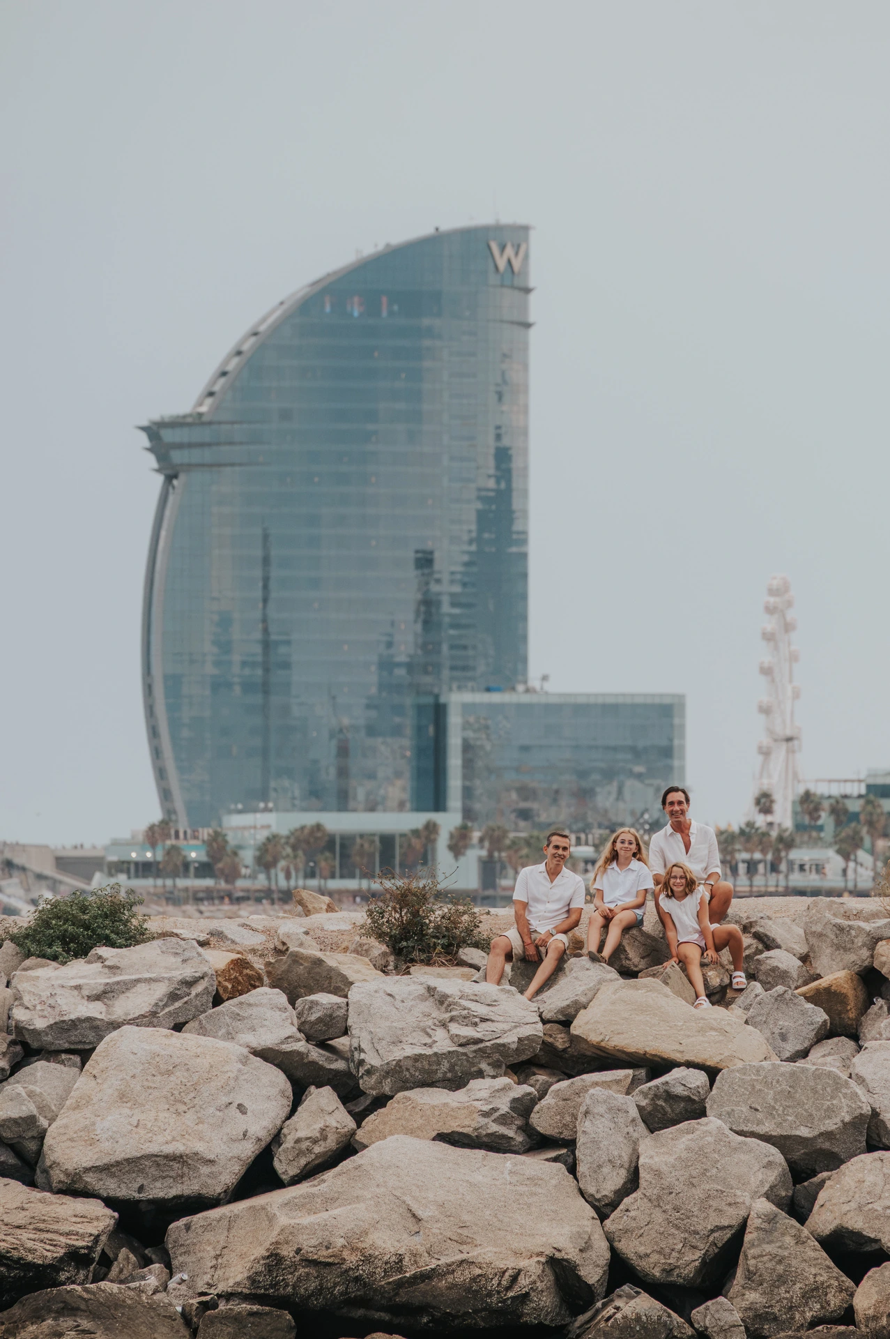 The family sitting together on the waterfront rocks, with the W hotel in the background during their family photoshoot in Barcelona.