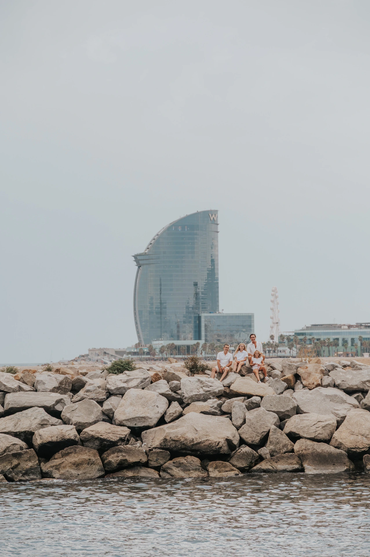 The family sitting together on the waterfront rocks, with the W hotel in the background during their family photoshoot in Barcelona.