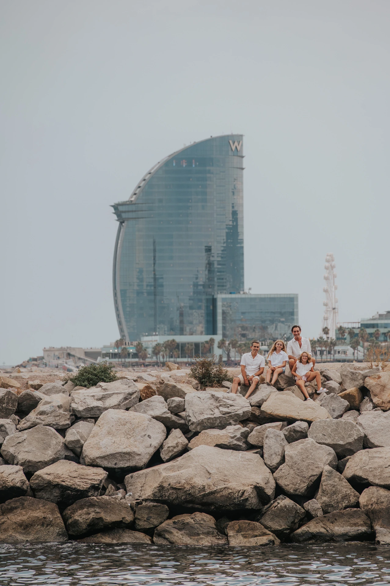 The family sitting together on the waterfront rocks, with the W hotel in the background during their family photoshoot in Barcelona.