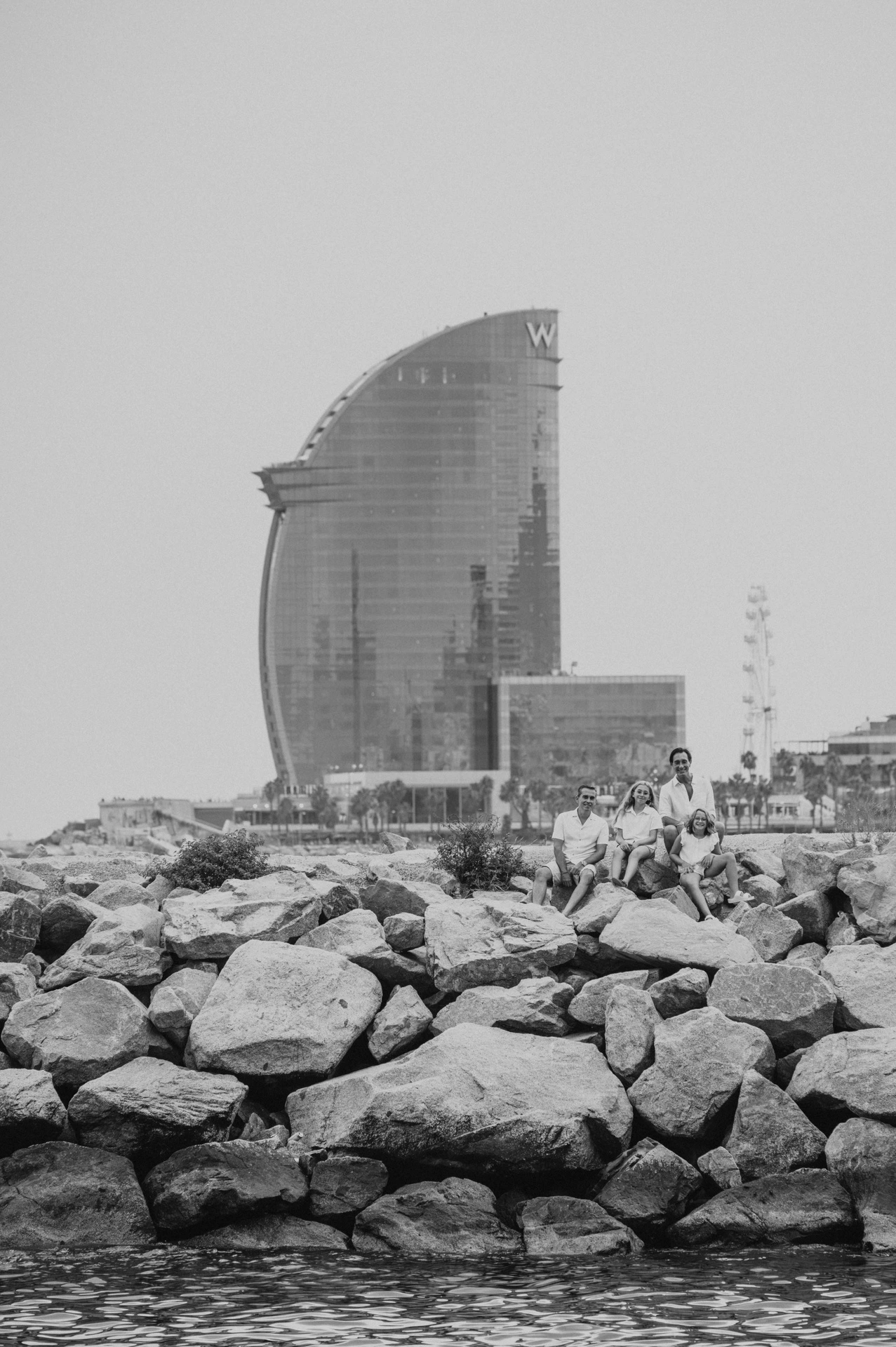 Black-and-white photograph: the family sitting together on the waterfront rocks, with the W hotel in the background during their family photoshoot in Barcelona.