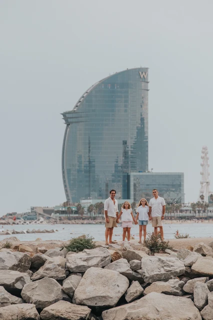 A waterfront family moment beside the W hotel | Barcelona family photoshoot The family together on the rocky waterfront with the W hotel rising behind them during their family photoshoot in Barcelona.