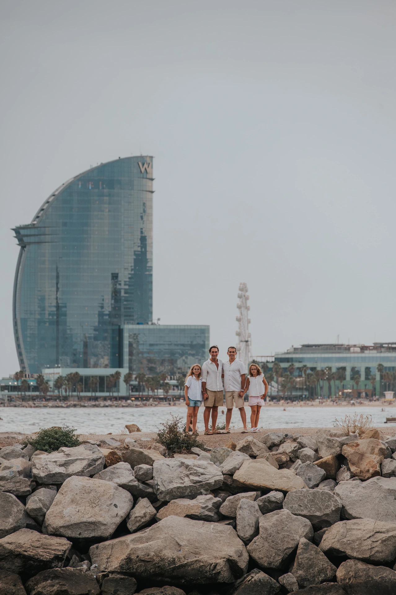 A wide view of the family standing on the rocky waterfront, with Barcelona’s sail-shaped W hotel behind them during their family photoshoot in Barcelona.