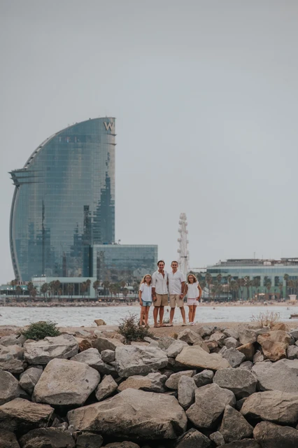 A wide view of the family standing on the rocky waterfront, with Barcelona’s sail-shaped W hotel behind them during their family photoshoot in Barcelona.