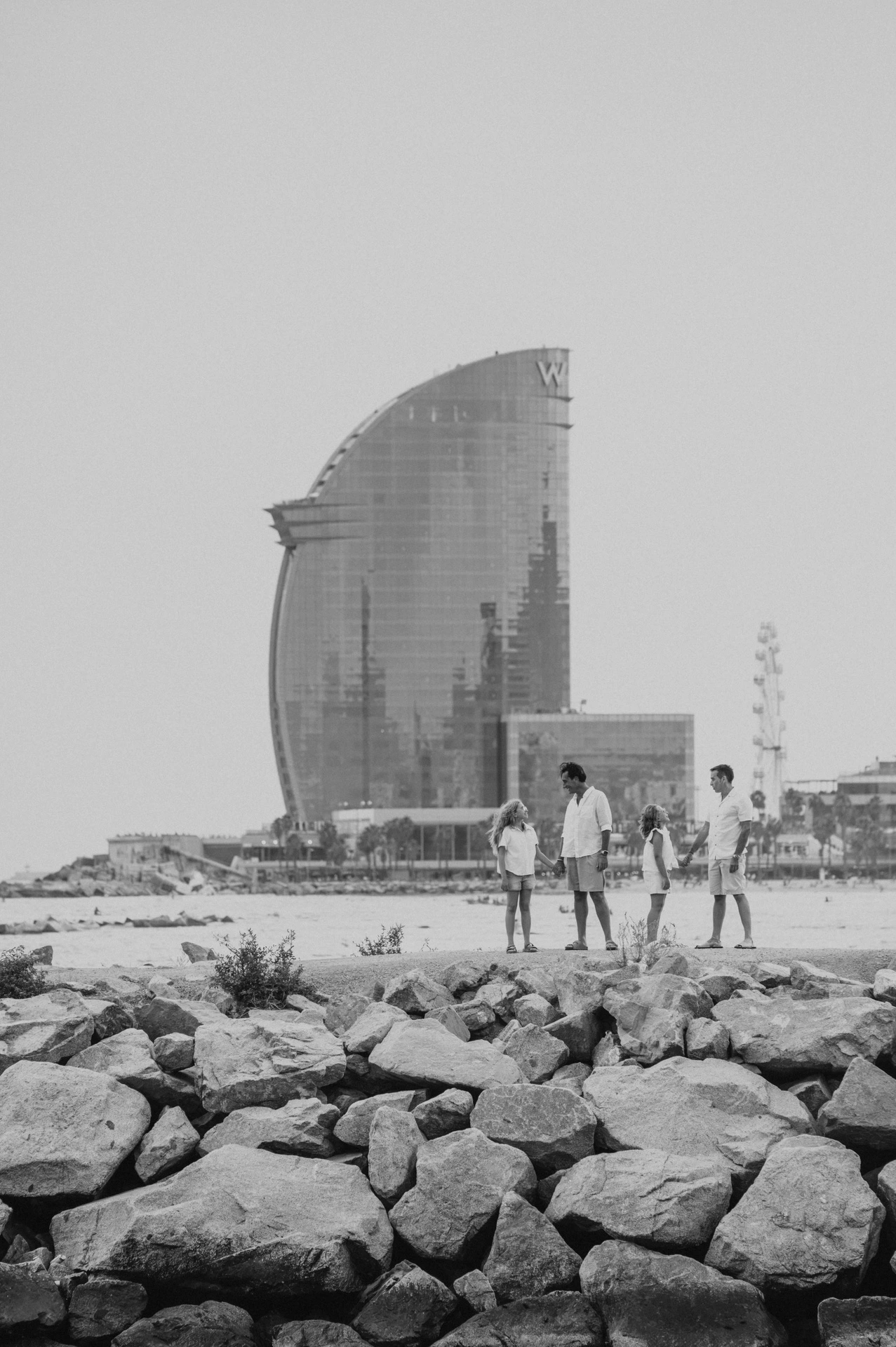 Black-and-white photograph: a wide view of the family standing on the rocky waterfront, with Barcelona’s sail-shaped W hotel behind them during their family photoshoot in Barcelona.
