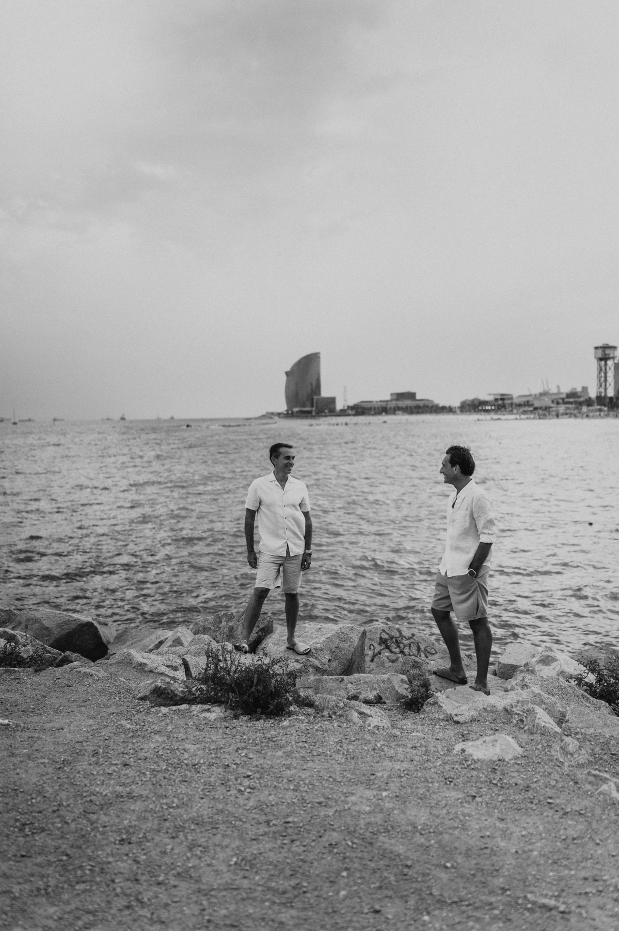 Black-and-white photograph: the two parents standing apart on the rocky shoreline and turning towards each other during their family photoshoot in Barcelona.