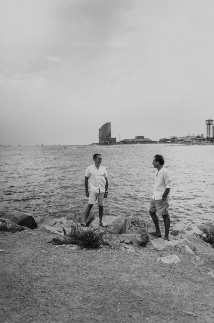 A moment for the parents beside the sea in black and white | Barcelona family photoshoot Black-and-white photograph: the two parents standing apart on the rocky shoreline and turning towards each other during their family photoshoot in Barcelona.