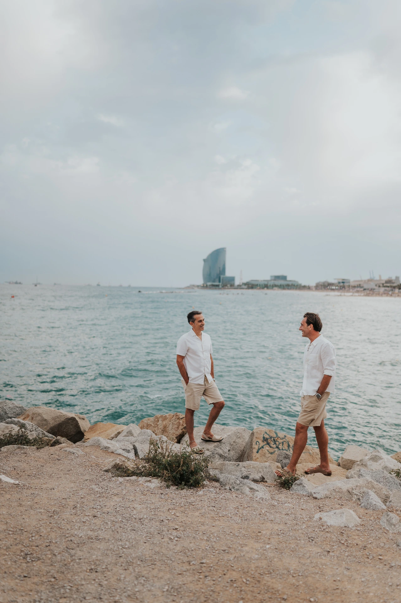 The two parents standing apart on the rocky shoreline and turning towards each other during their family photoshoot in Barcelona.