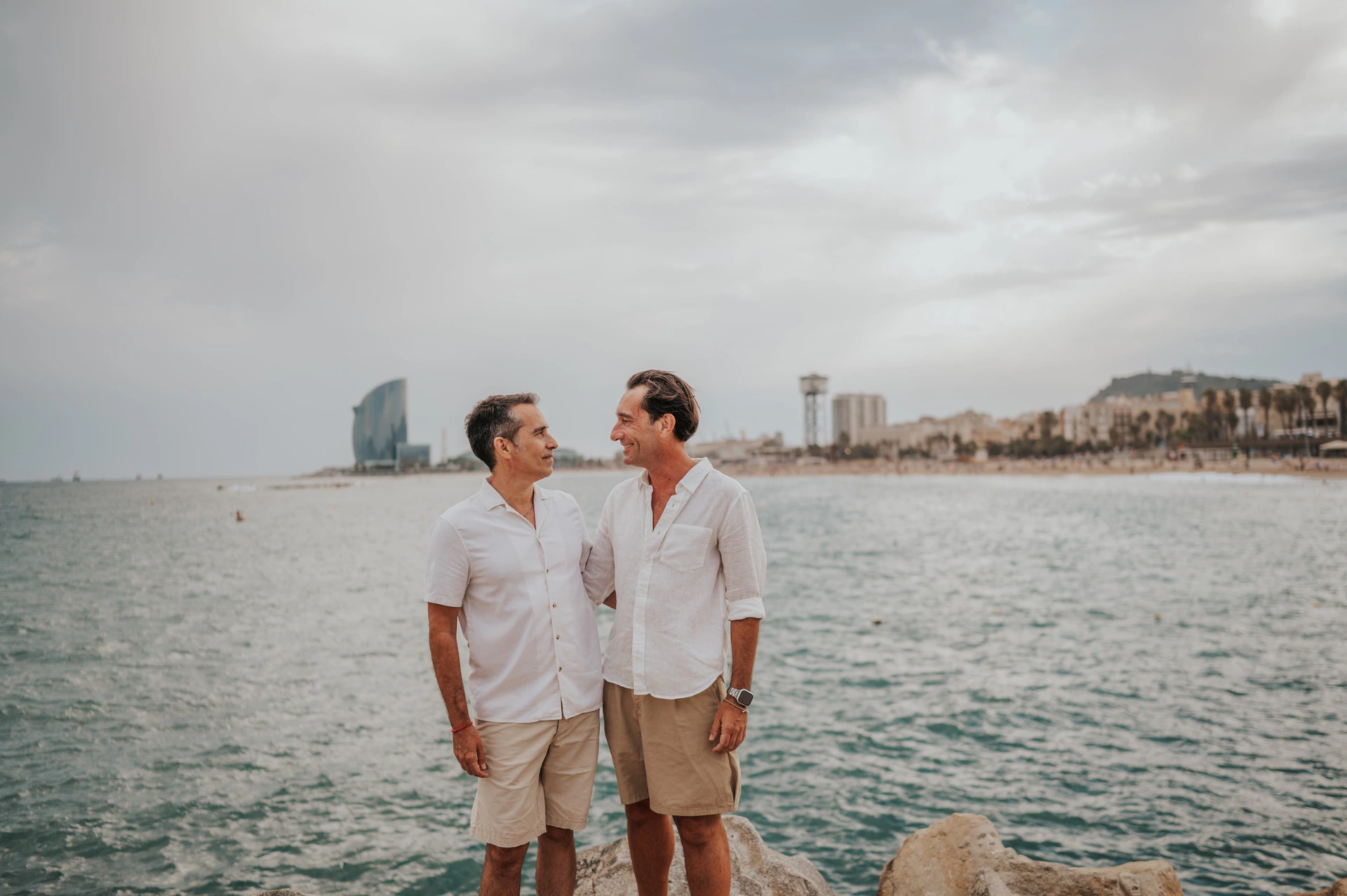 The two parents standing together beside the water and rocky shoreline during their family photoshoot in Barcelona.