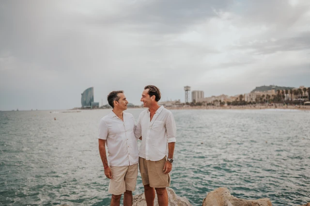 The two parents standing together beside the water and rocky shoreline during their family photoshoot in Barcelona.