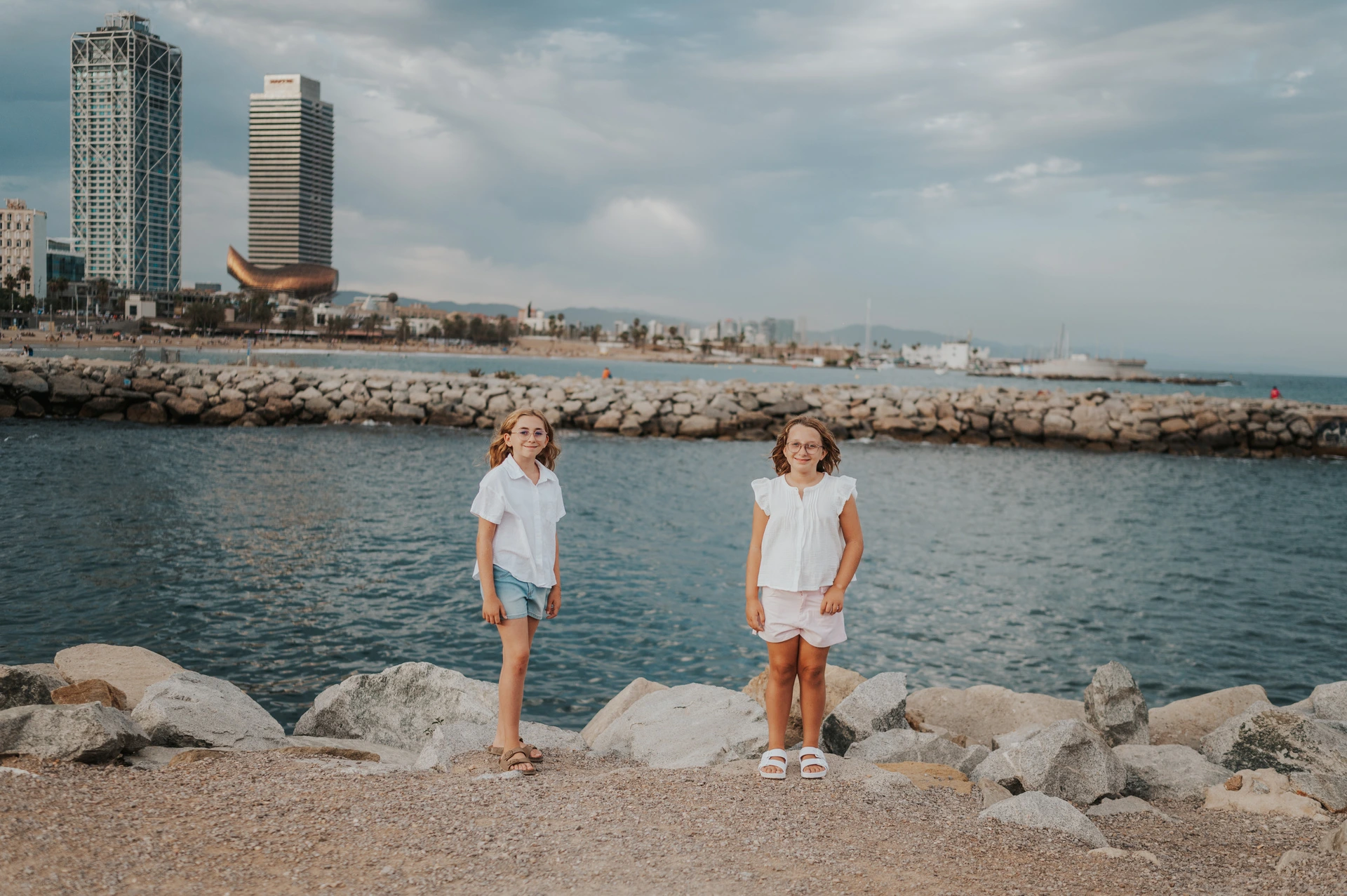 The two sisters standing apart on the waterfront path beside the rocks and sea during their family photoshoot in Barcelona.