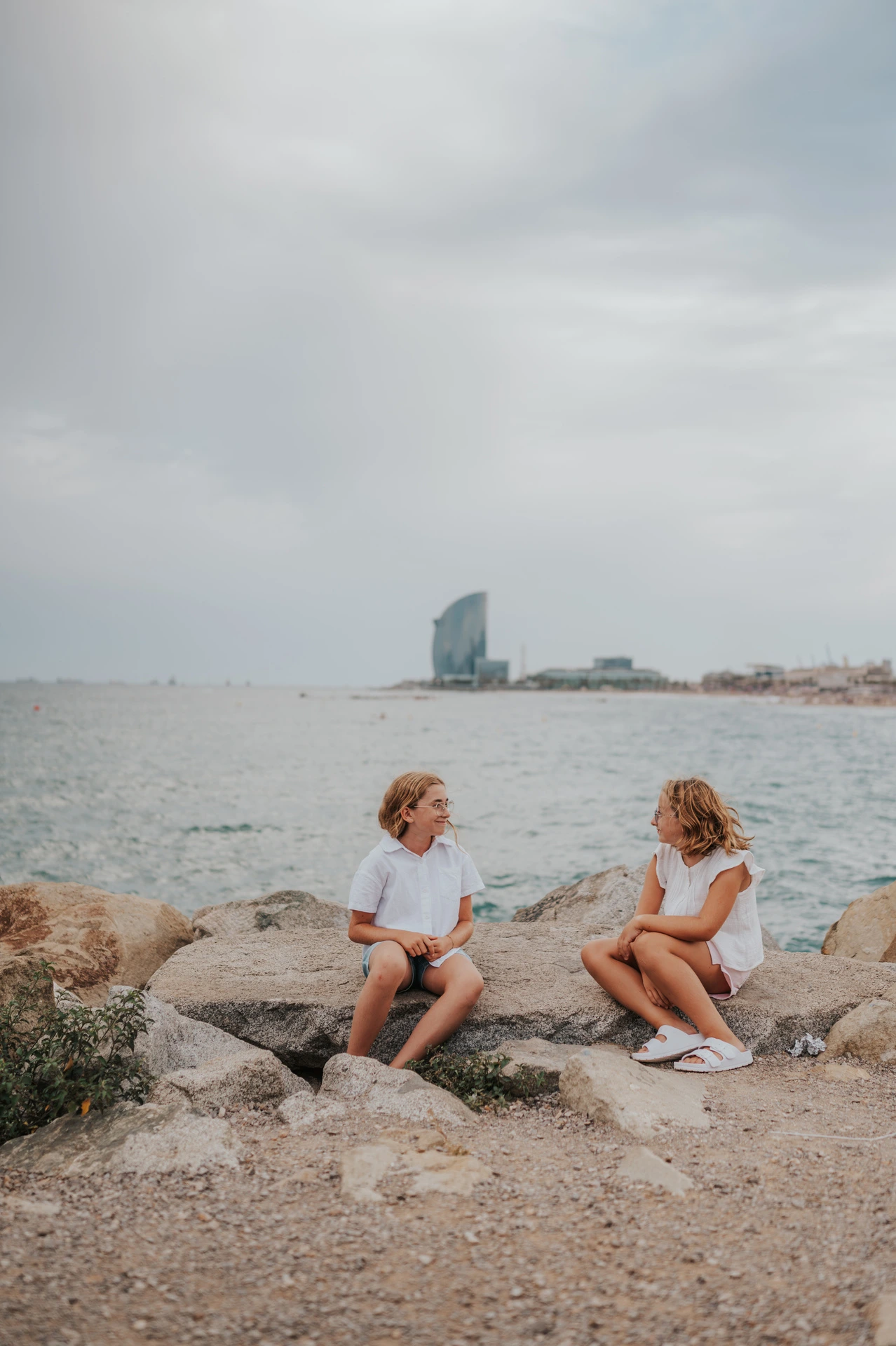 The two sisters sitting on the rocks beside the sea and turning towards each other during their family photoshoot in Barcelona.