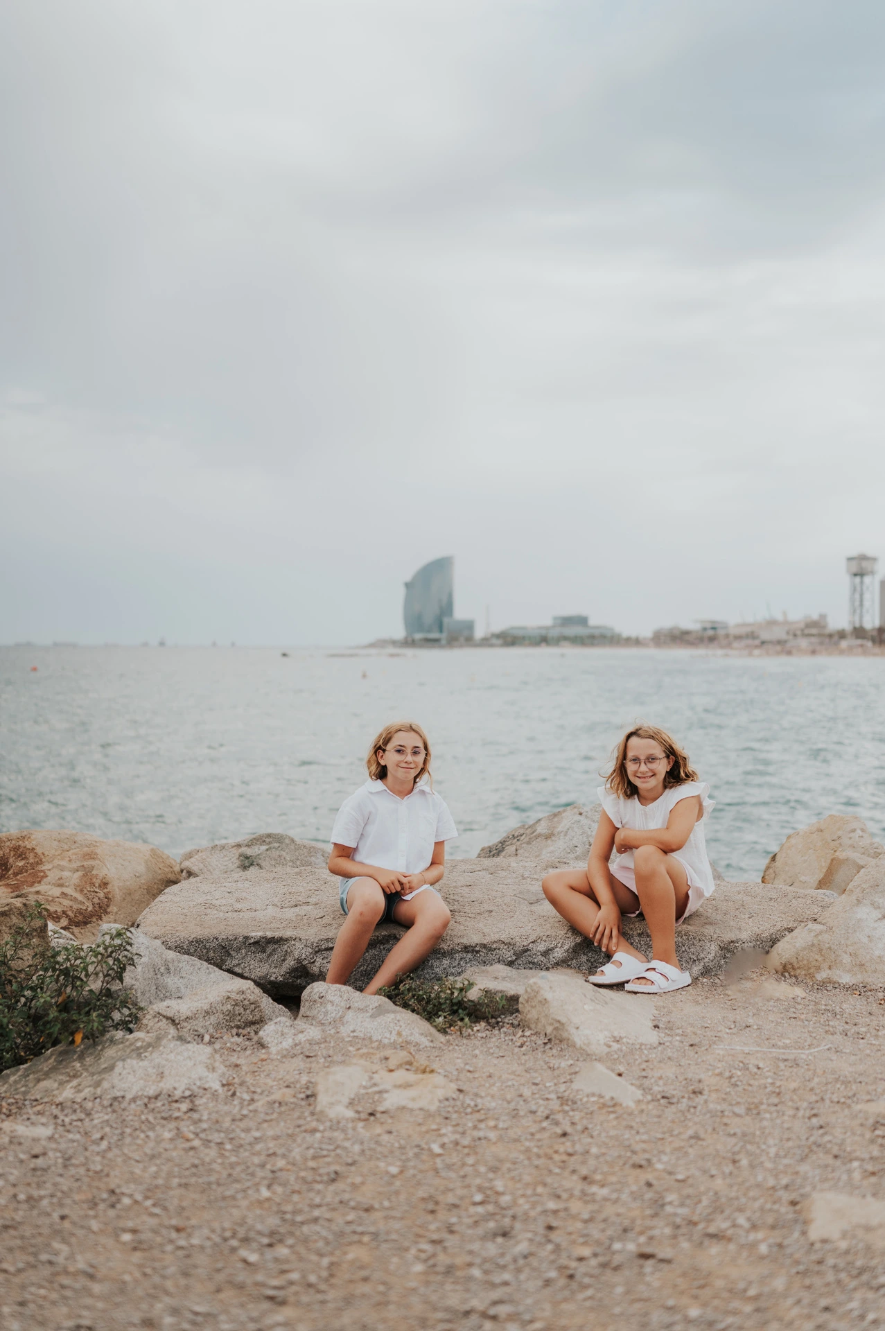 The two sisters sitting on the rocks beside the sea and turning towards each other during their family photoshoot in Barcelona.