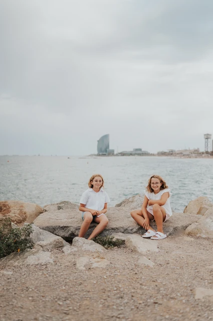 The sisters sitting beside the sea | Barcelona family photoshoot The two sisters sitting on the rocks beside the sea and turning towards each other during their family photoshoot in Barcelona.