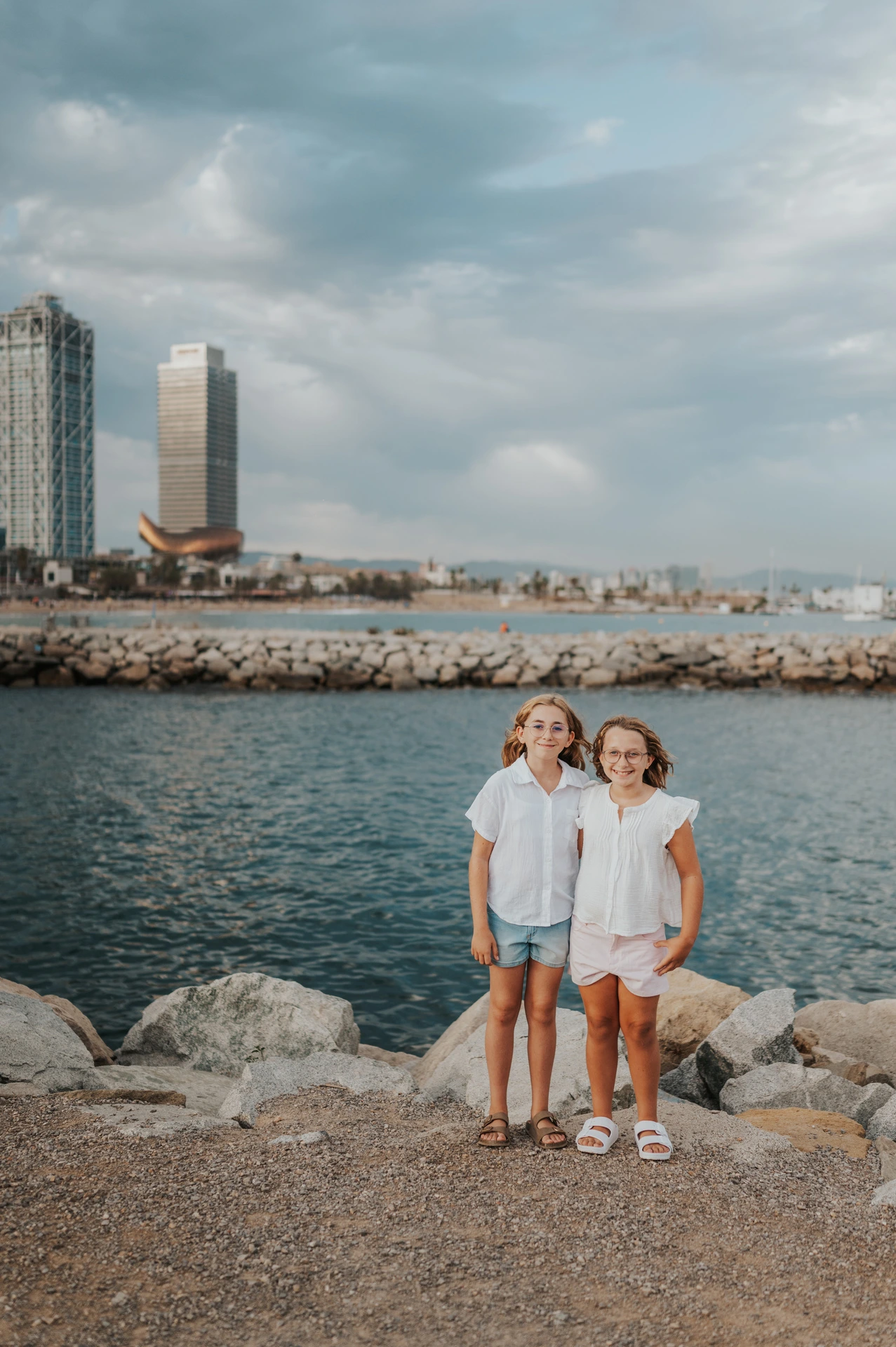 The two sisters standing together beside the water, with Barcelona’s waterfront buildings behind them during their family photoshoot in Barcelona.