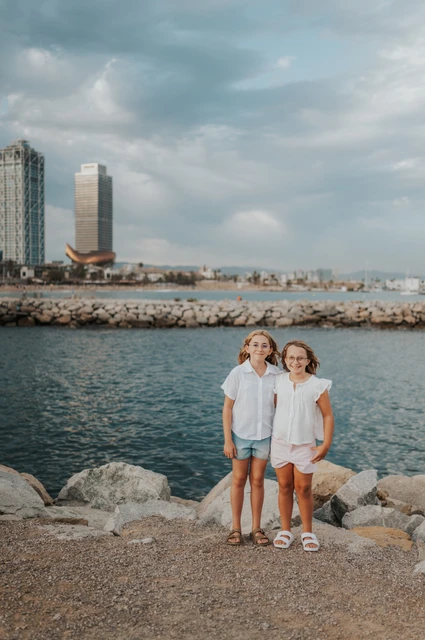 Sister portraits beside the water | Barcelona family photoshoot The two sisters standing together beside the water, with Barcelona’s waterfront buildings behind them during their family photoshoot in Barcelona.