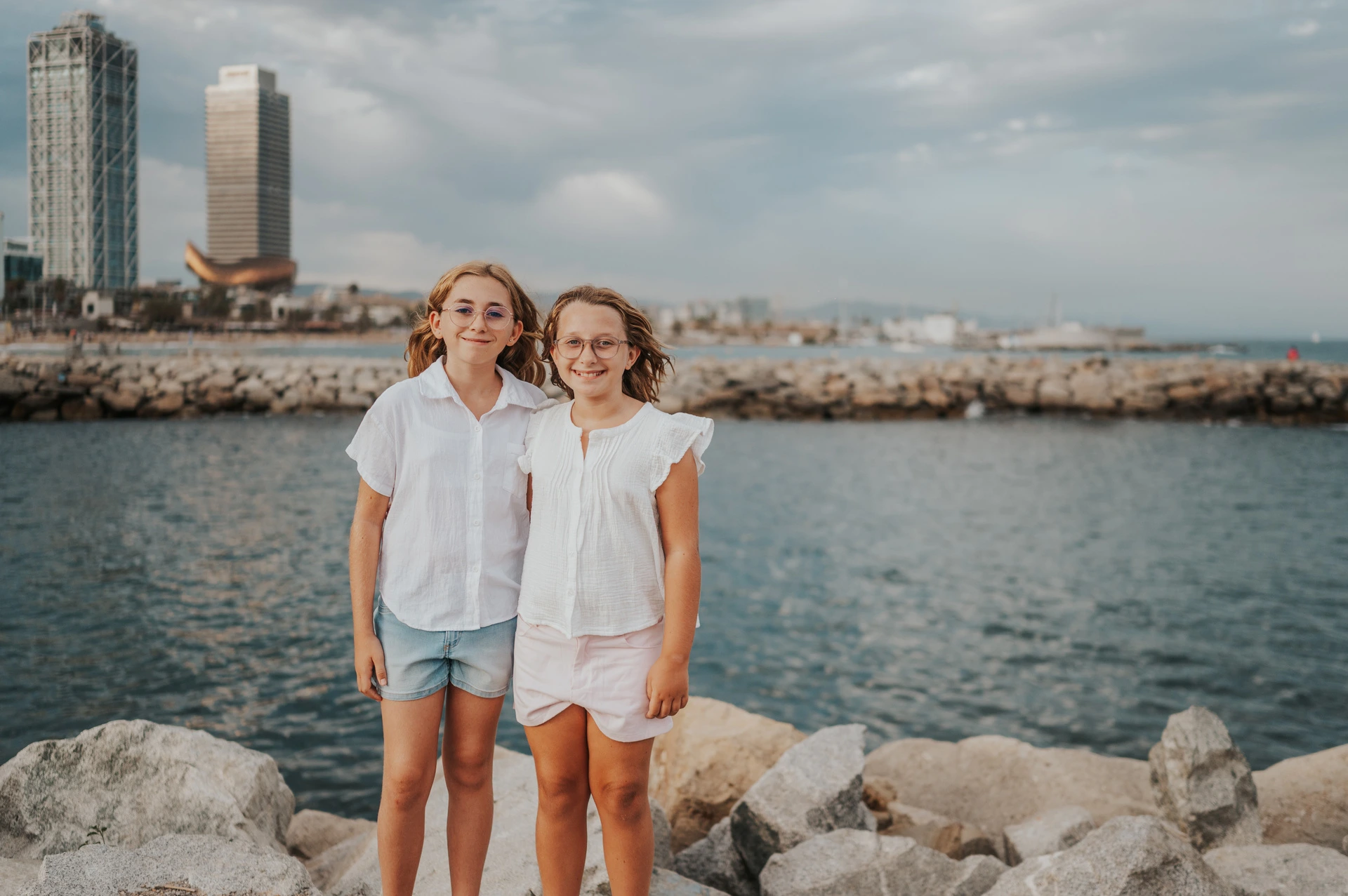 The two sisters standing together beside the water, with Barcelona’s waterfront buildings behind them during their family photoshoot in Barcelona.