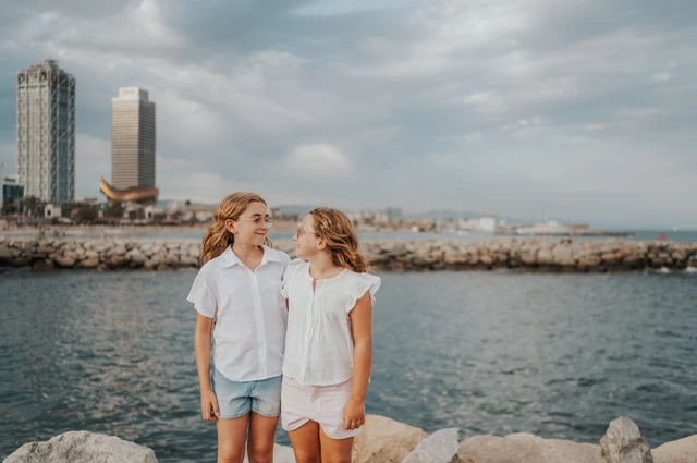 The two sisters standing together beside the water, with Barcelona’s waterfront buildings behind them during their family photoshoot in Barcelona.