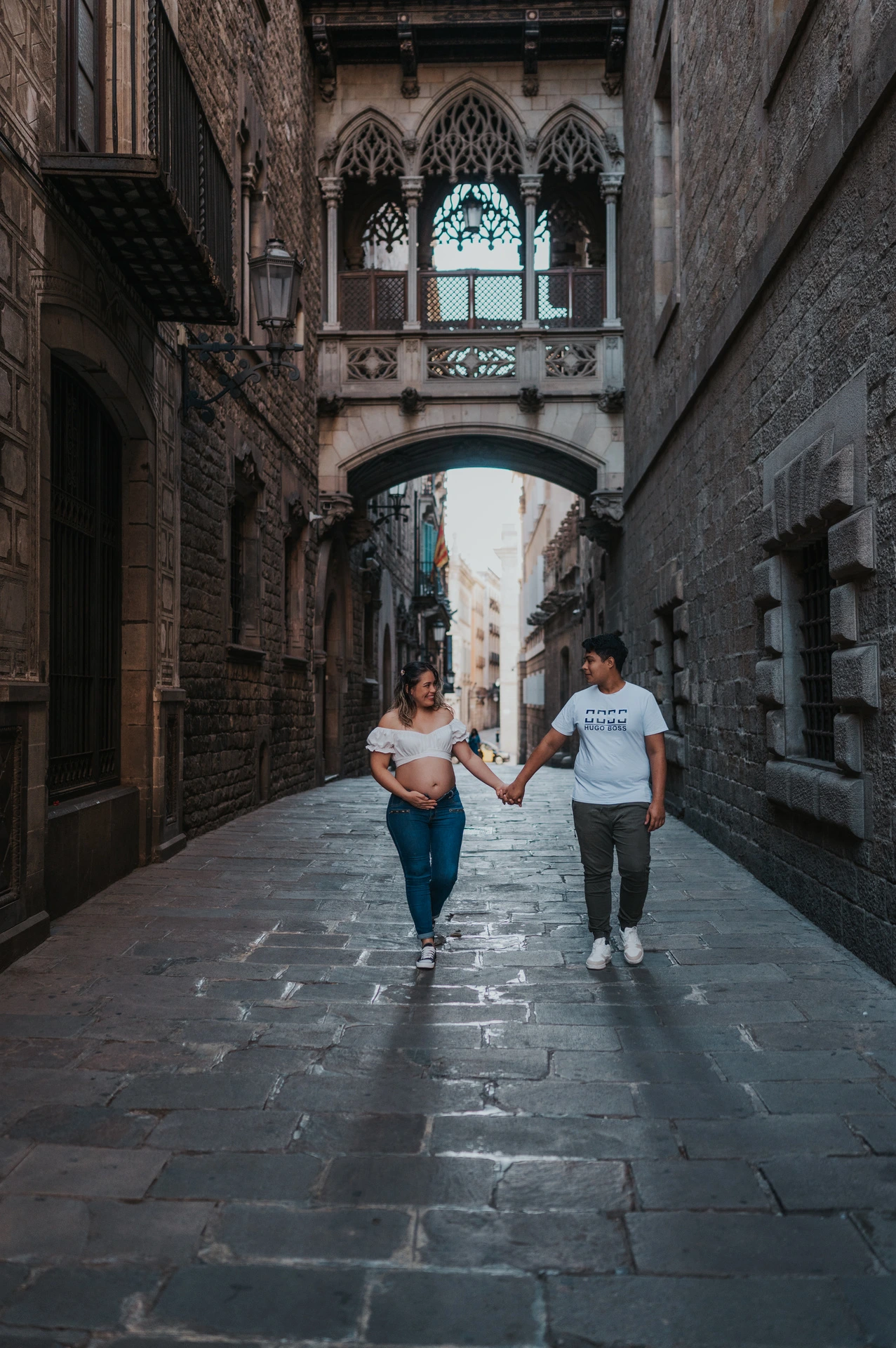 The couple walking hand in hand along the stone street beneath the ornate bridge during their maternity photoshoot in Barcelona.