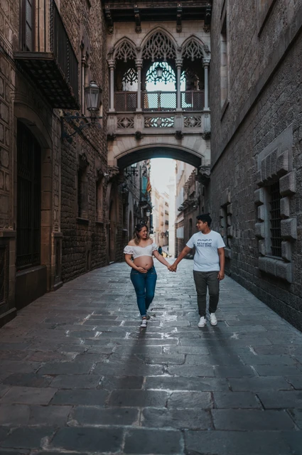 A hand-in-hand walk beneath the bridge | Barcelona maternity photoshoot The couple walking hand in hand along the stone street beneath the ornate bridge during their maternity photoshoot in Barcelona.