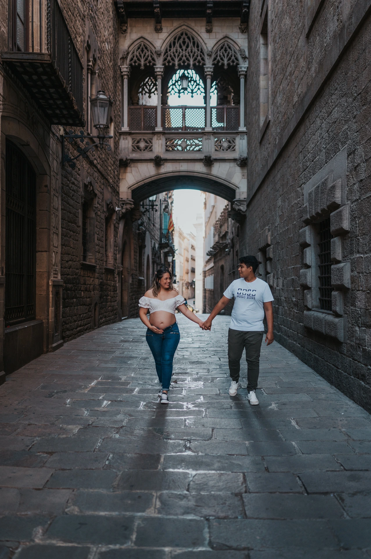 The couple walking hand in hand along the stone street beneath the ornate bridge during their maternity photoshoot in Barcelona.