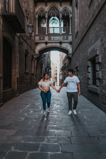 A hand-in-hand walk beneath the bridge | Barcelona maternity photoshoot The couple walking hand in hand along the stone street beneath the ornate bridge during their maternity photoshoot in Barcelona.
