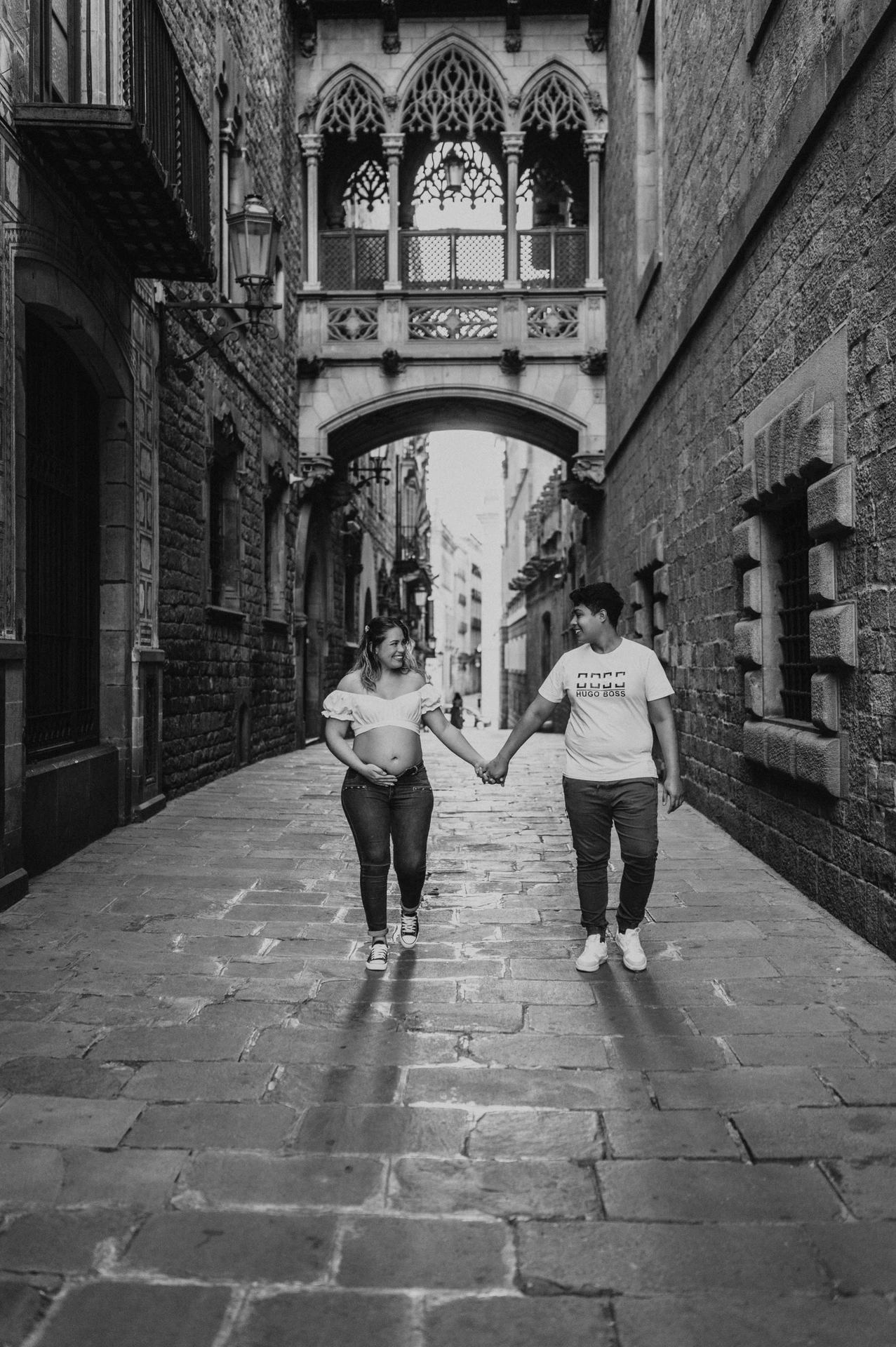 Black-and-white photograph: the couple walking hand in hand along the stone street beneath the ornate bridge during their maternity photoshoot in Barcelona.