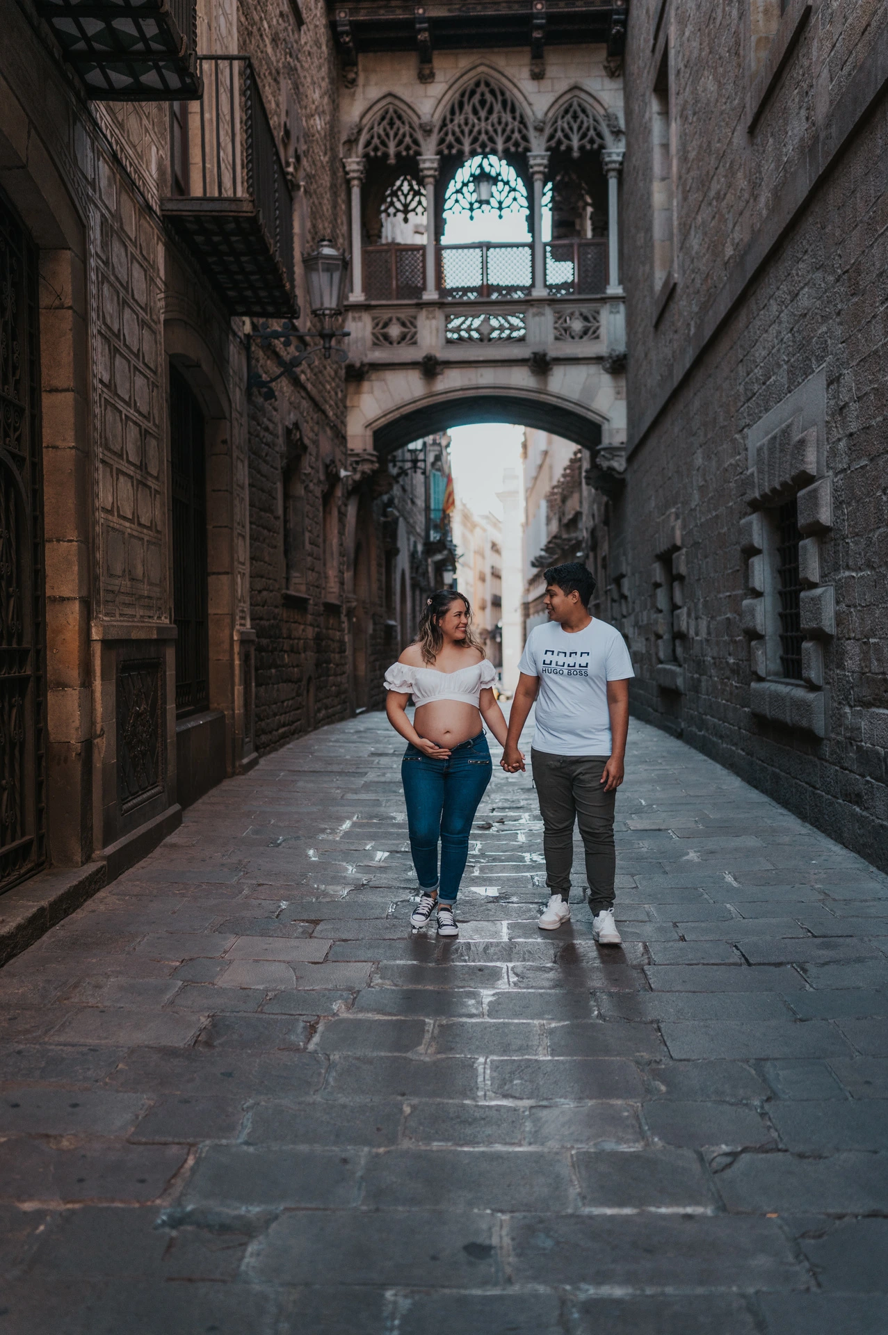 The couple walking hand in hand along the stone street beneath the ornate bridge during their maternity photoshoot in Barcelona.