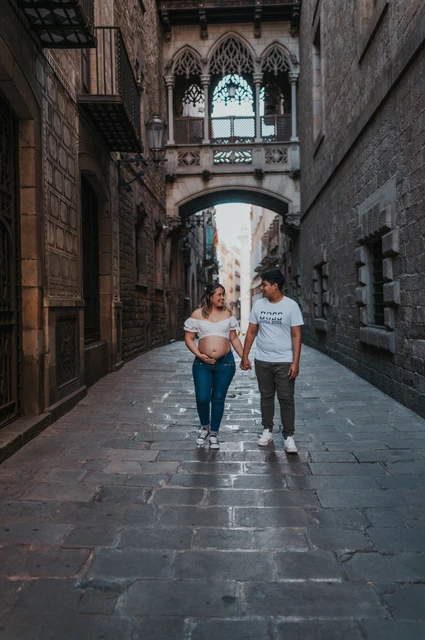 A hand-in-hand walk beneath the bridge | Barcelona maternity photoshoot The couple walking hand in hand along the stone street beneath the ornate bridge during their maternity photoshoot in Barcelona.