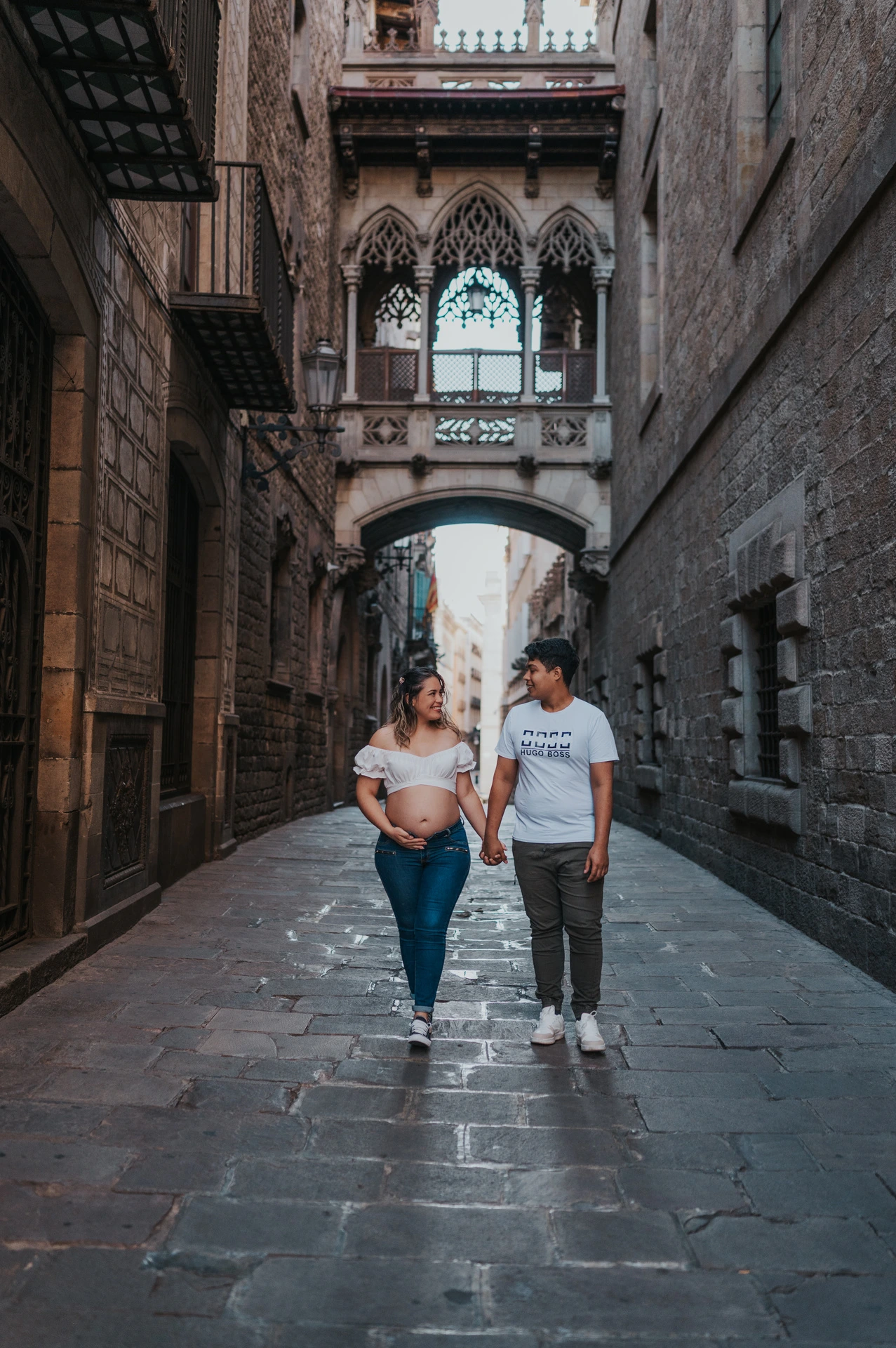 The couple walking hand in hand along the stone street beneath the ornate bridge during their maternity photoshoot in Barcelona.