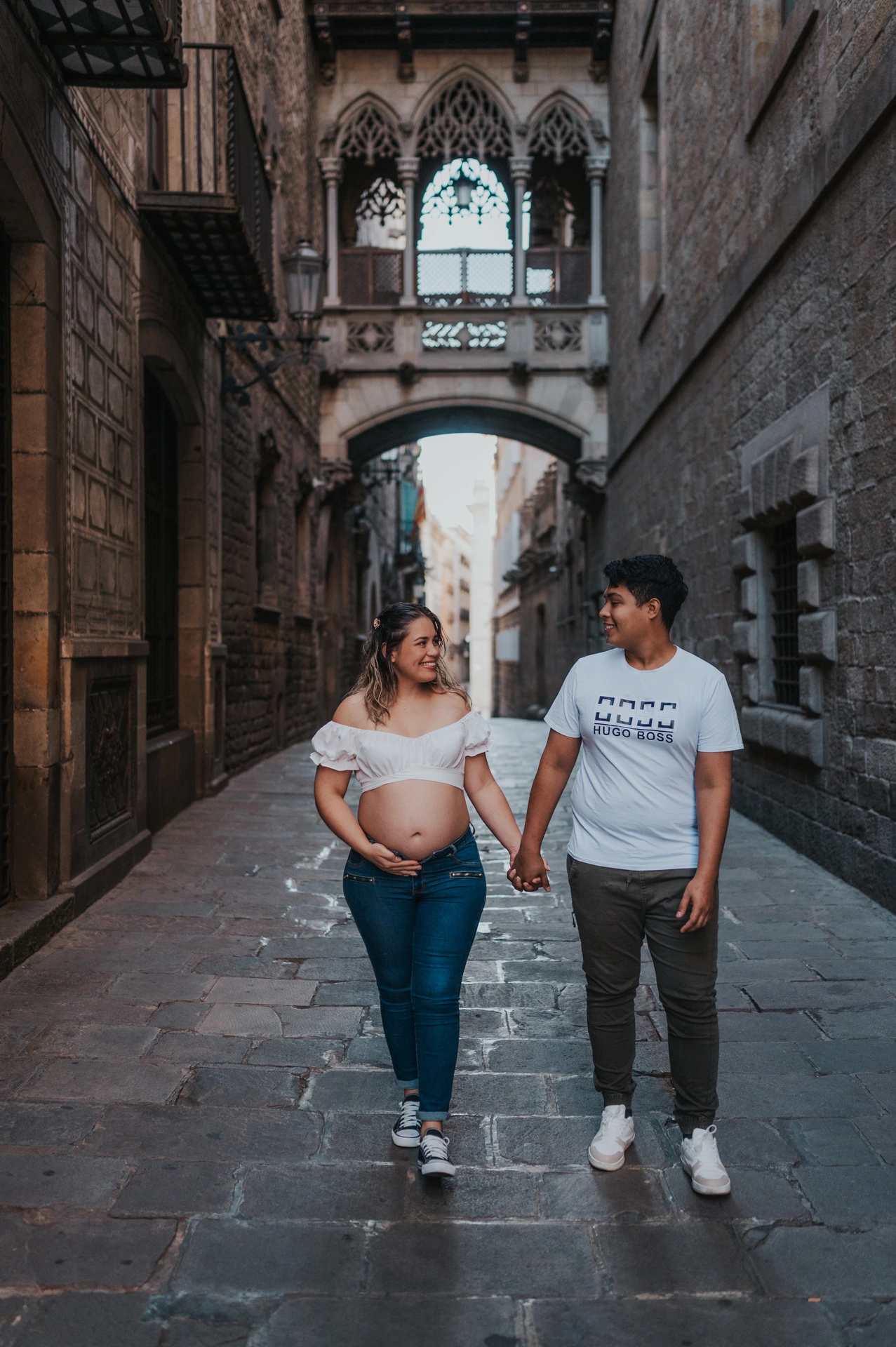 The couple walking hand in hand along the stone street beneath the ornate bridge during their maternity photoshoot in Barcelona.