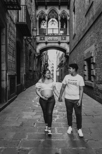A hand-in-hand walk beneath the bridge in black and white | Barcelona maternity photoshoot Black-and-white photograph: the couple walking hand in hand along the stone street beneath the ornate bridge during their maternity photoshoot in Barcelona.