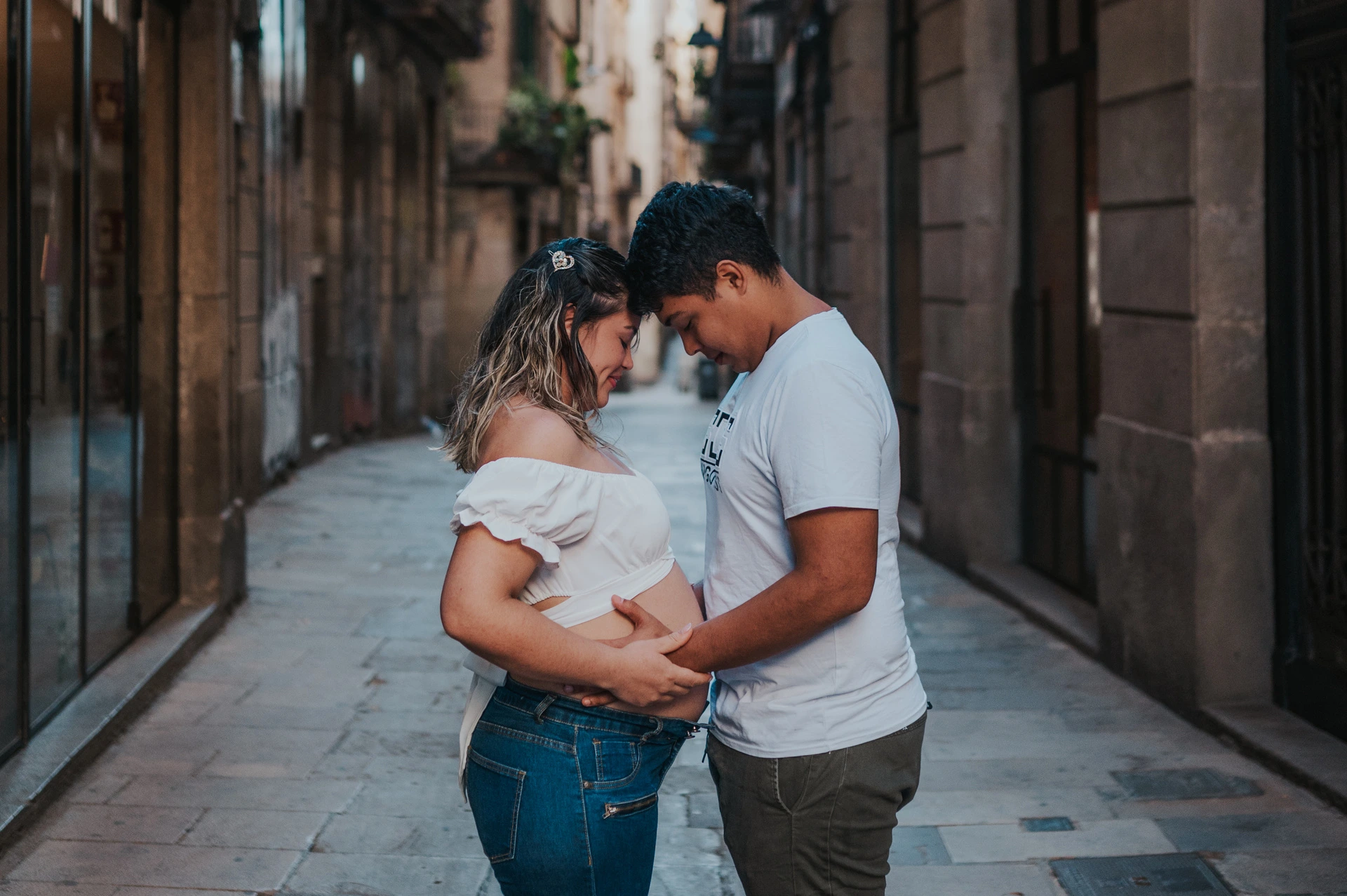 The couple facing one another and holding her bump in the narrow street during their maternity photoshoot in Barcelona.