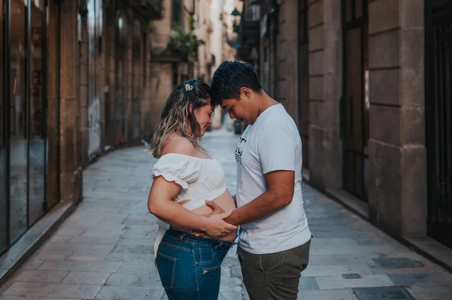 The couple facing one another and holding her bump in the narrow street during their maternity photoshoot in Barcelona.