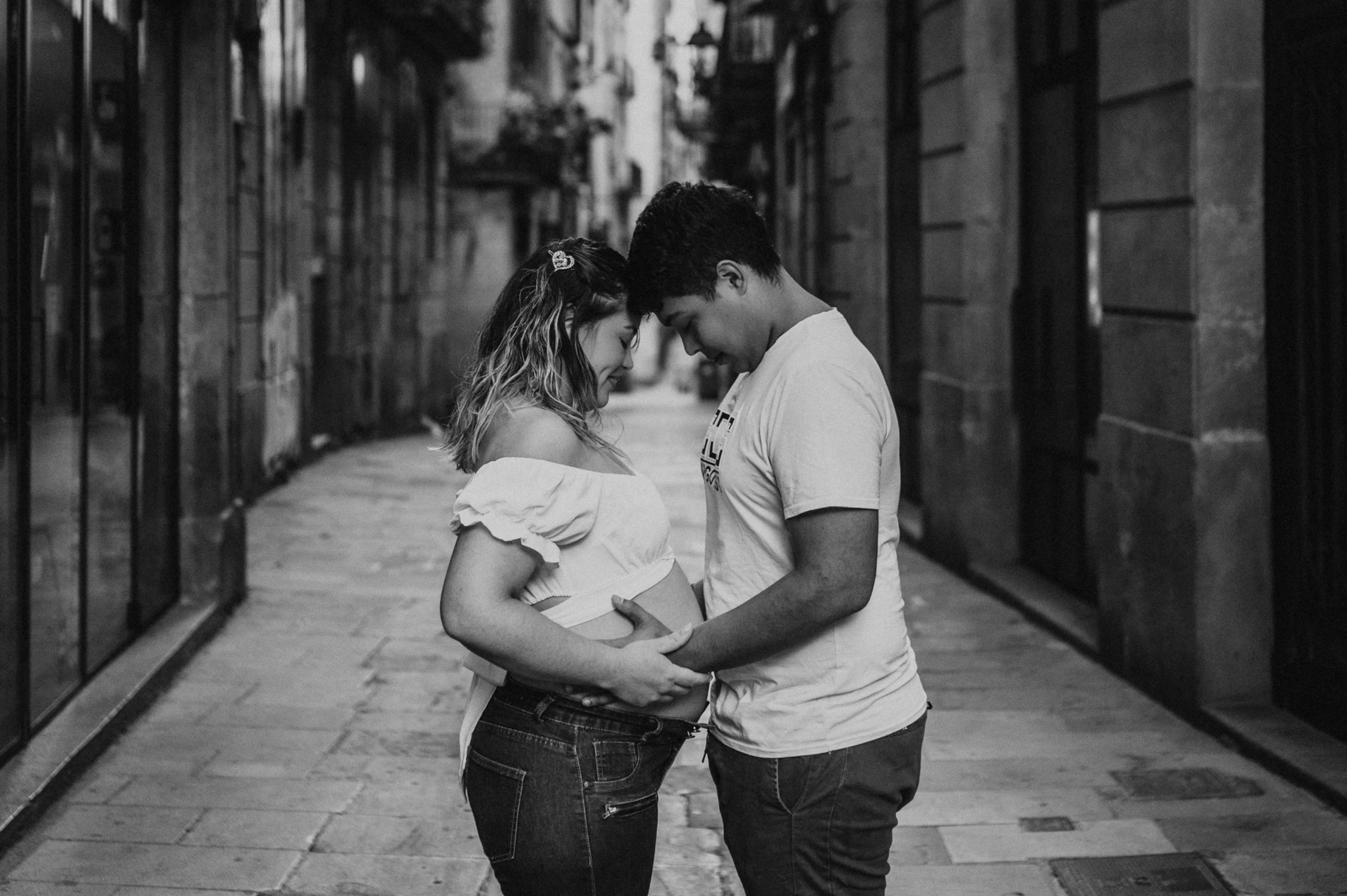 Black-and-white photograph: the couple facing one another and holding her bump in the narrow street during their maternity photoshoot in Barcelona.
