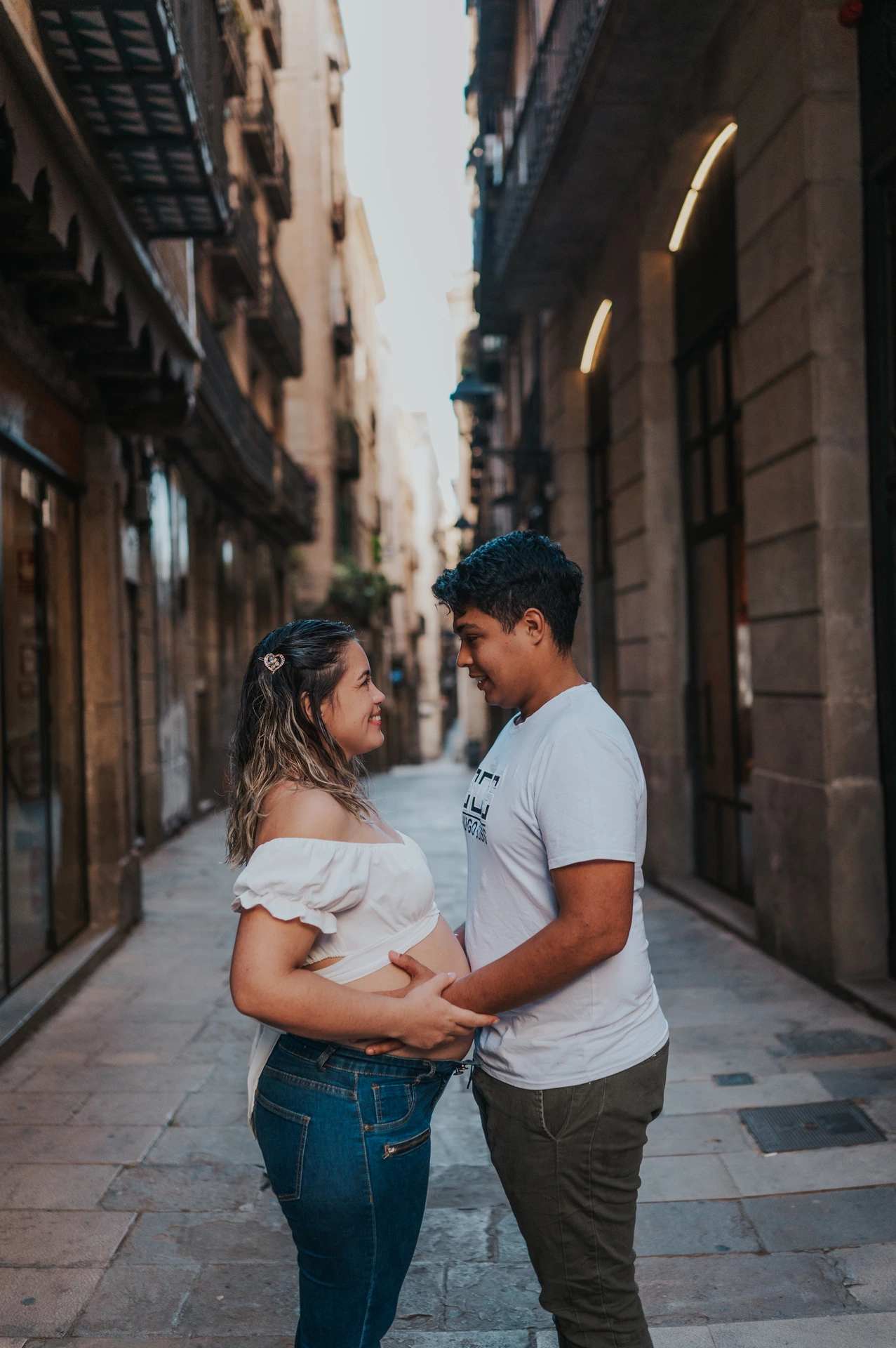 The couple facing one another and holding her bump in the narrow street during their maternity photoshoot in Barcelona.
