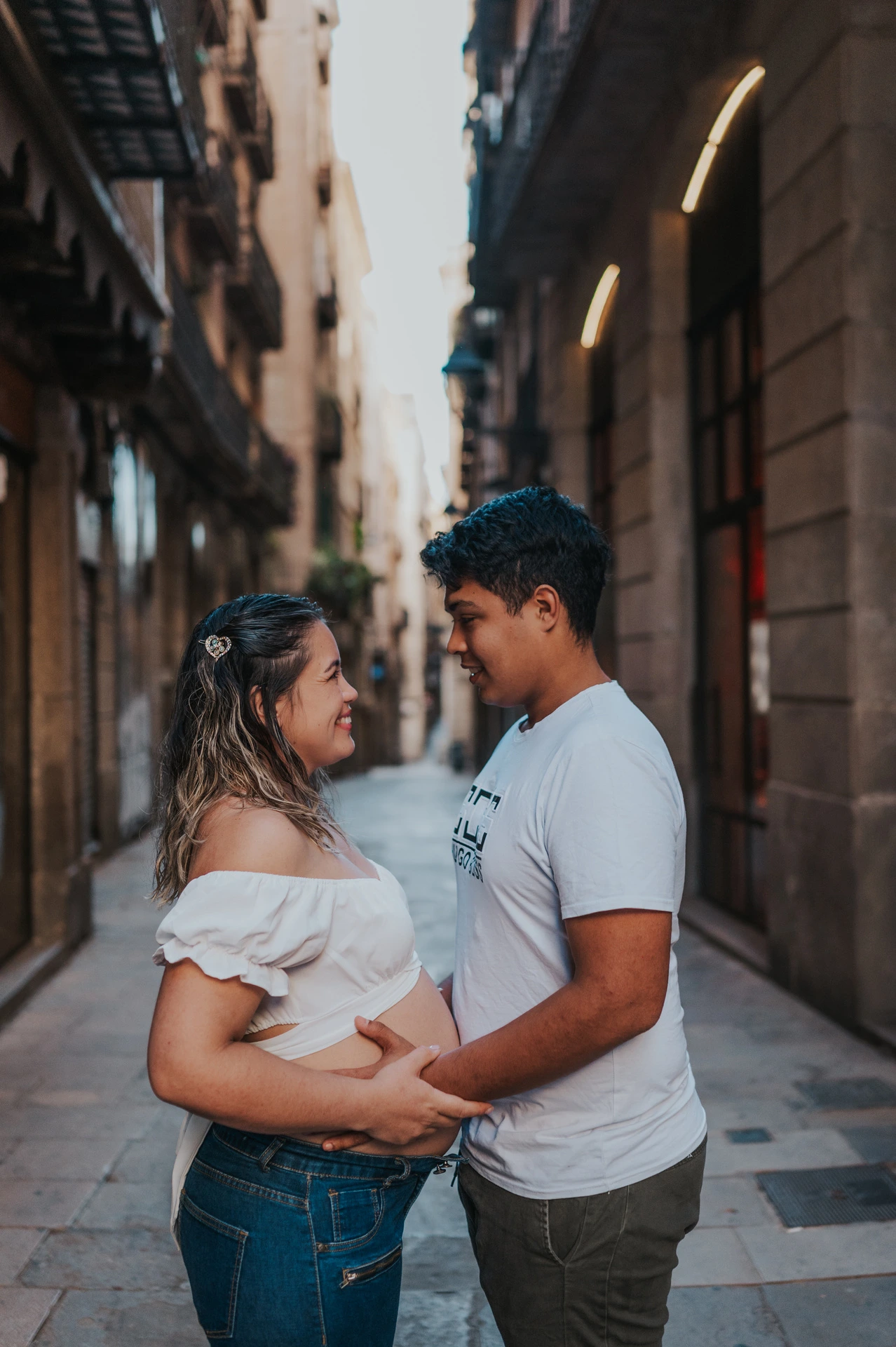 The couple facing one another and holding her bump in the narrow street during their maternity photoshoot in Barcelona.