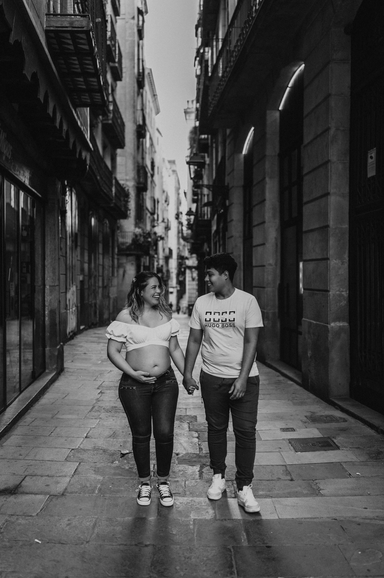 Black-and-white photograph: the couple walking together along a narrow street lined with old buildings during their maternity photoshoot in Barcelona.
