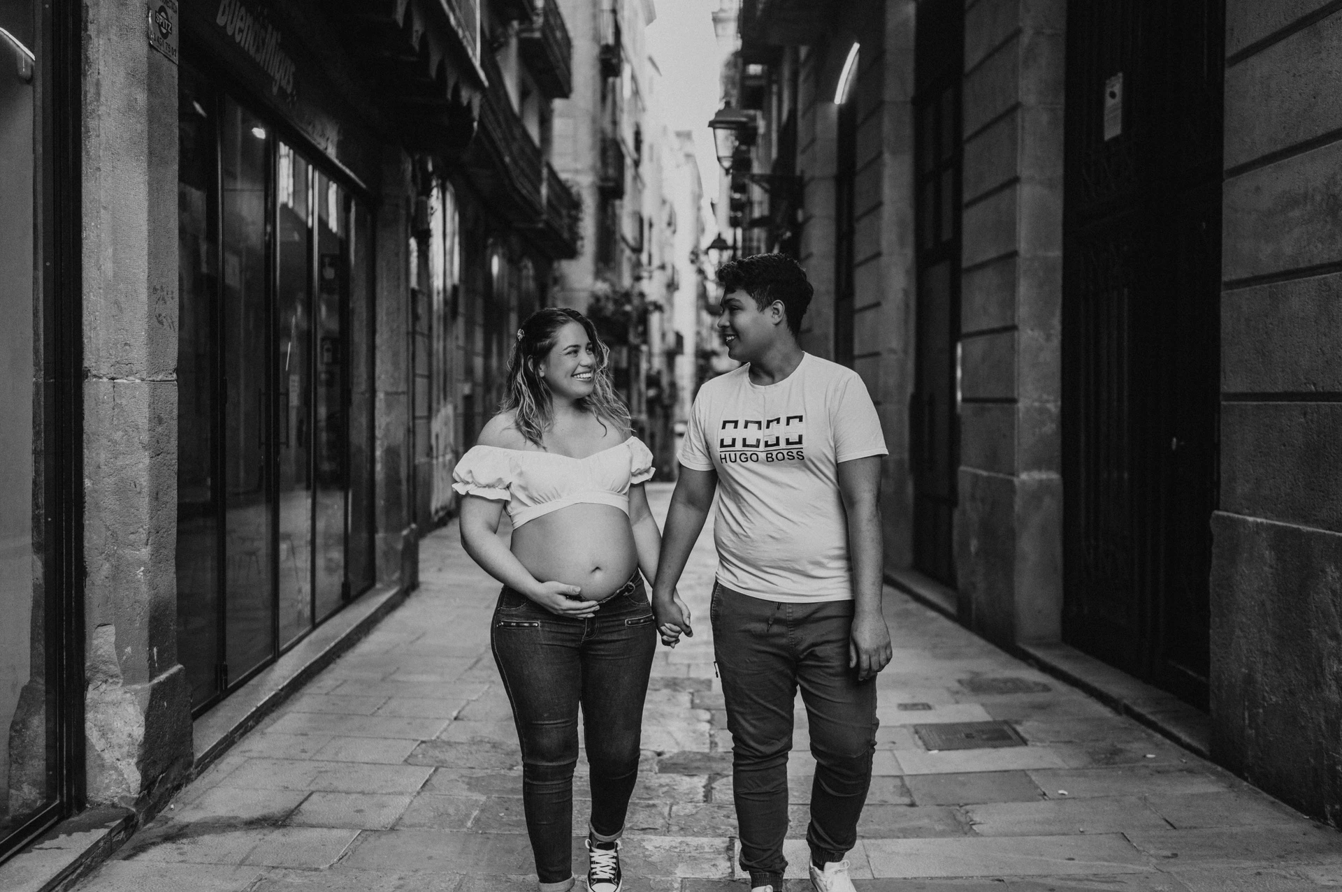 Black-and-white photograph: the couple walking together along a narrow street lined with old buildings during their maternity photoshoot in Barcelona.