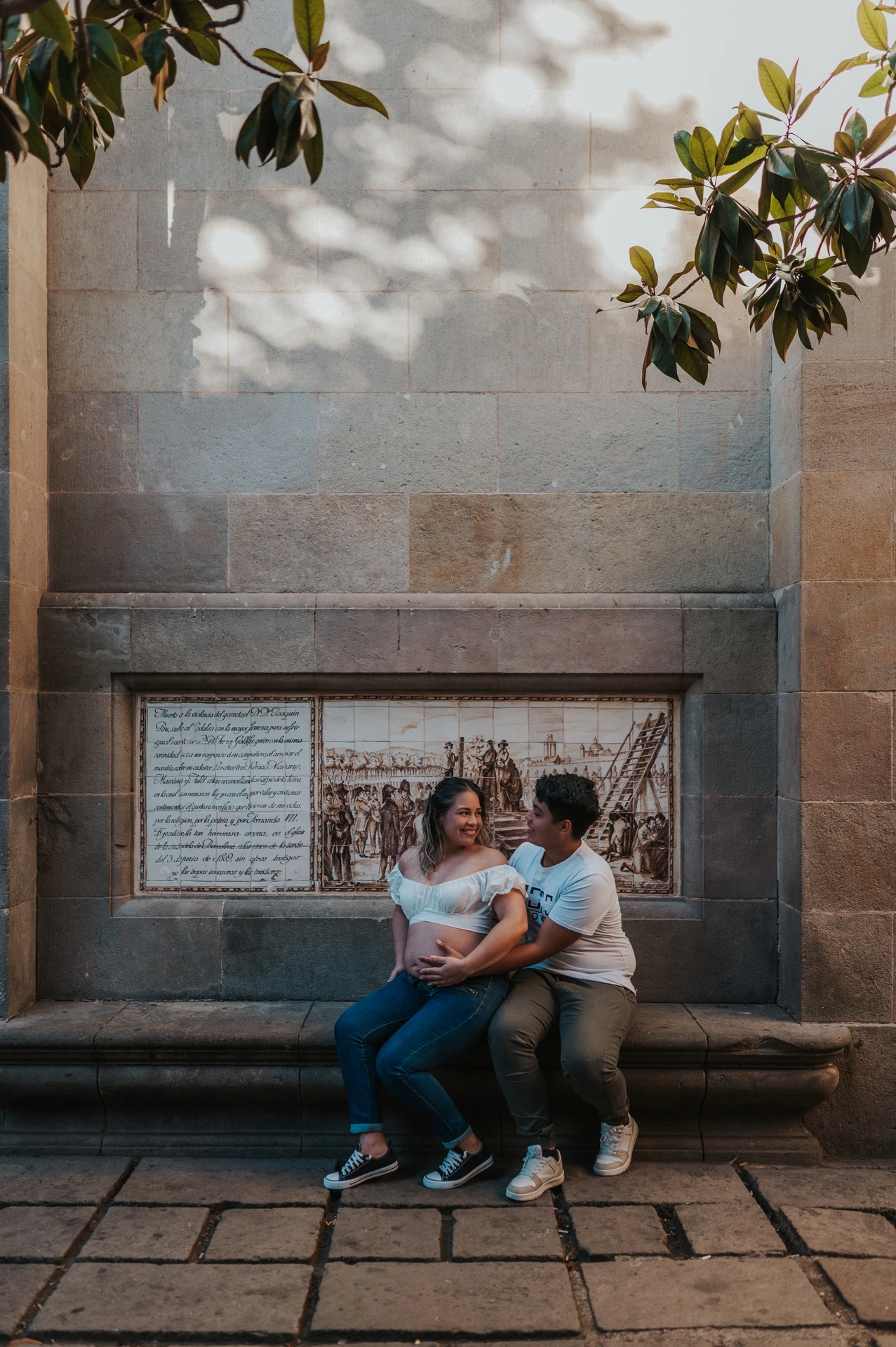 The couple sitting close together on a stone bench beneath a tree during their maternity photoshoot in Barcelona.
