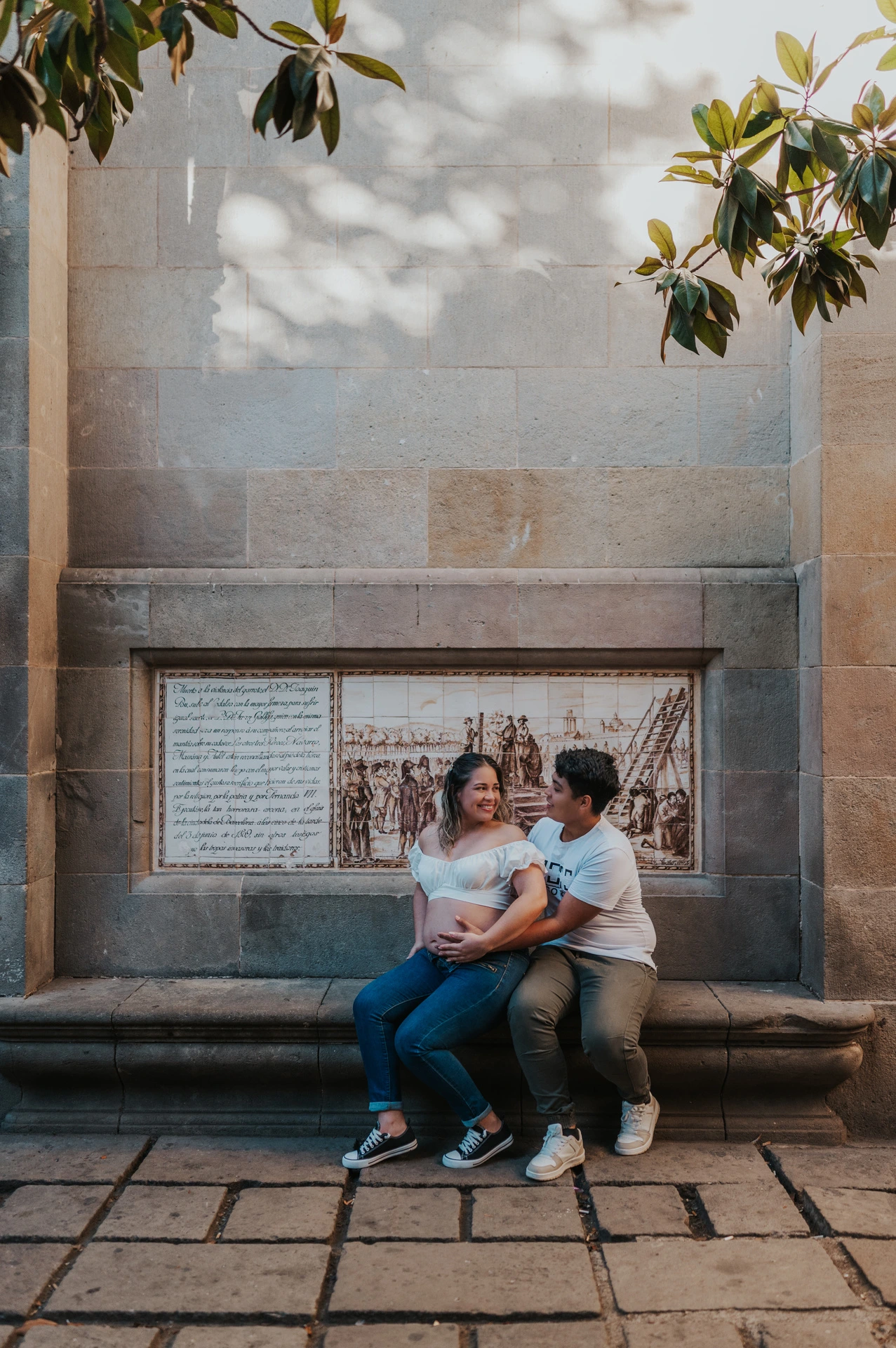 The couple sitting close together on a stone bench beneath a tree during their maternity photoshoot in Barcelona.