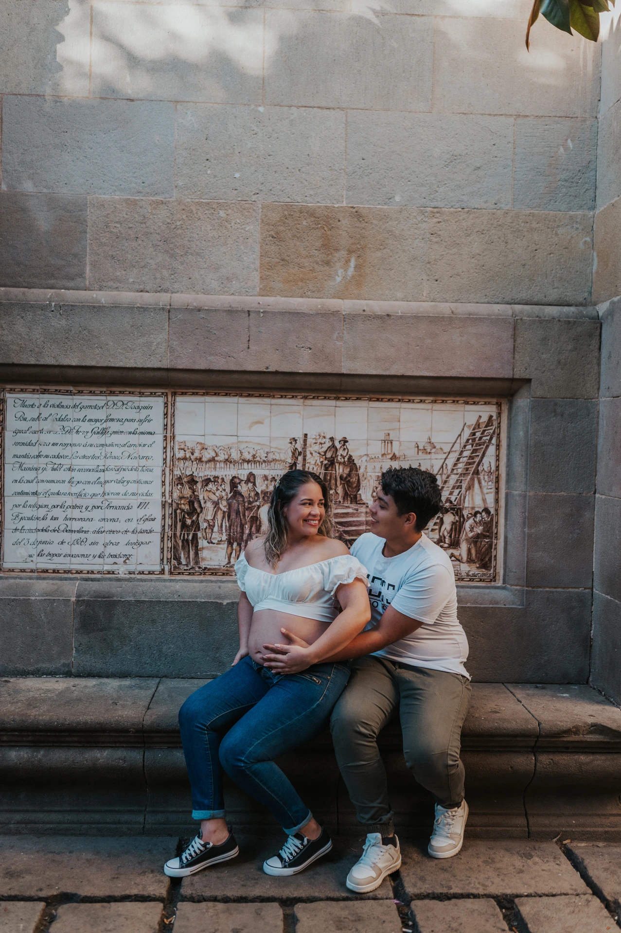 The couple sitting close together on a stone bench beneath a tree during their maternity photoshoot in Barcelona.