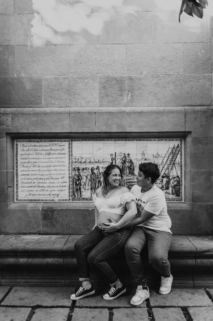 A moment together on the stone bench in black and white | Barcelona maternity photoshoot Black-and-white photograph: the couple sitting close together on a stone bench beneath a tree during their maternity photoshoot in Barcelona.