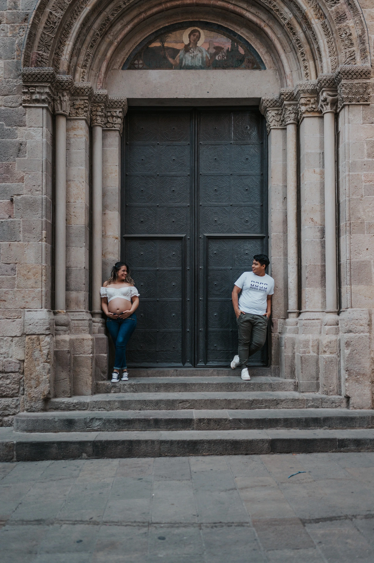 The couple standing on either side of a large doorway framed by carved stone and steps during their maternity photoshoot in Barcelona.