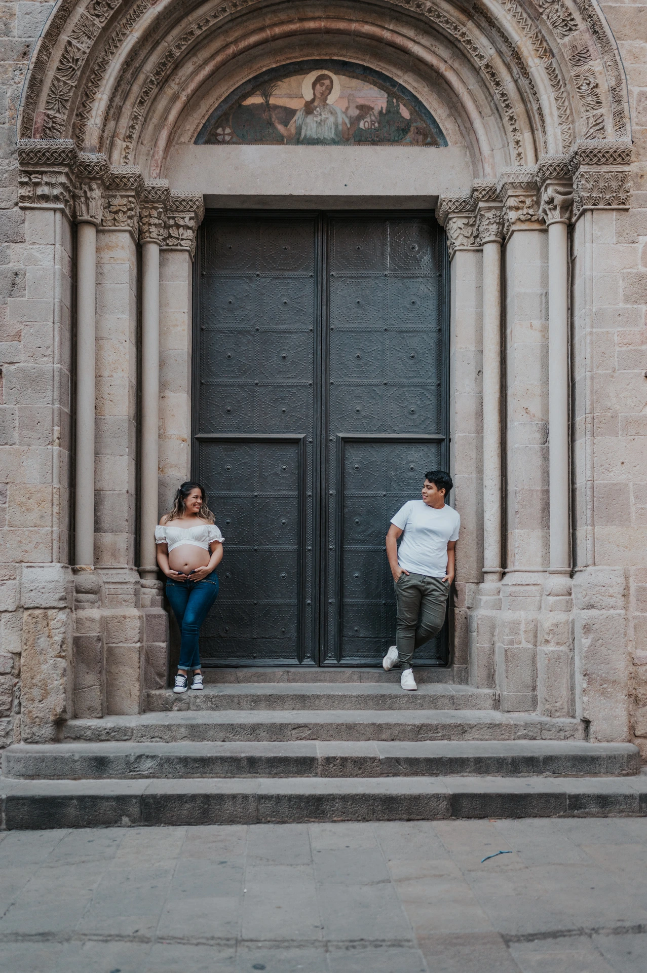 The couple standing on either side of a large doorway framed by carved stone and steps during their maternity photoshoot in Barcelona.