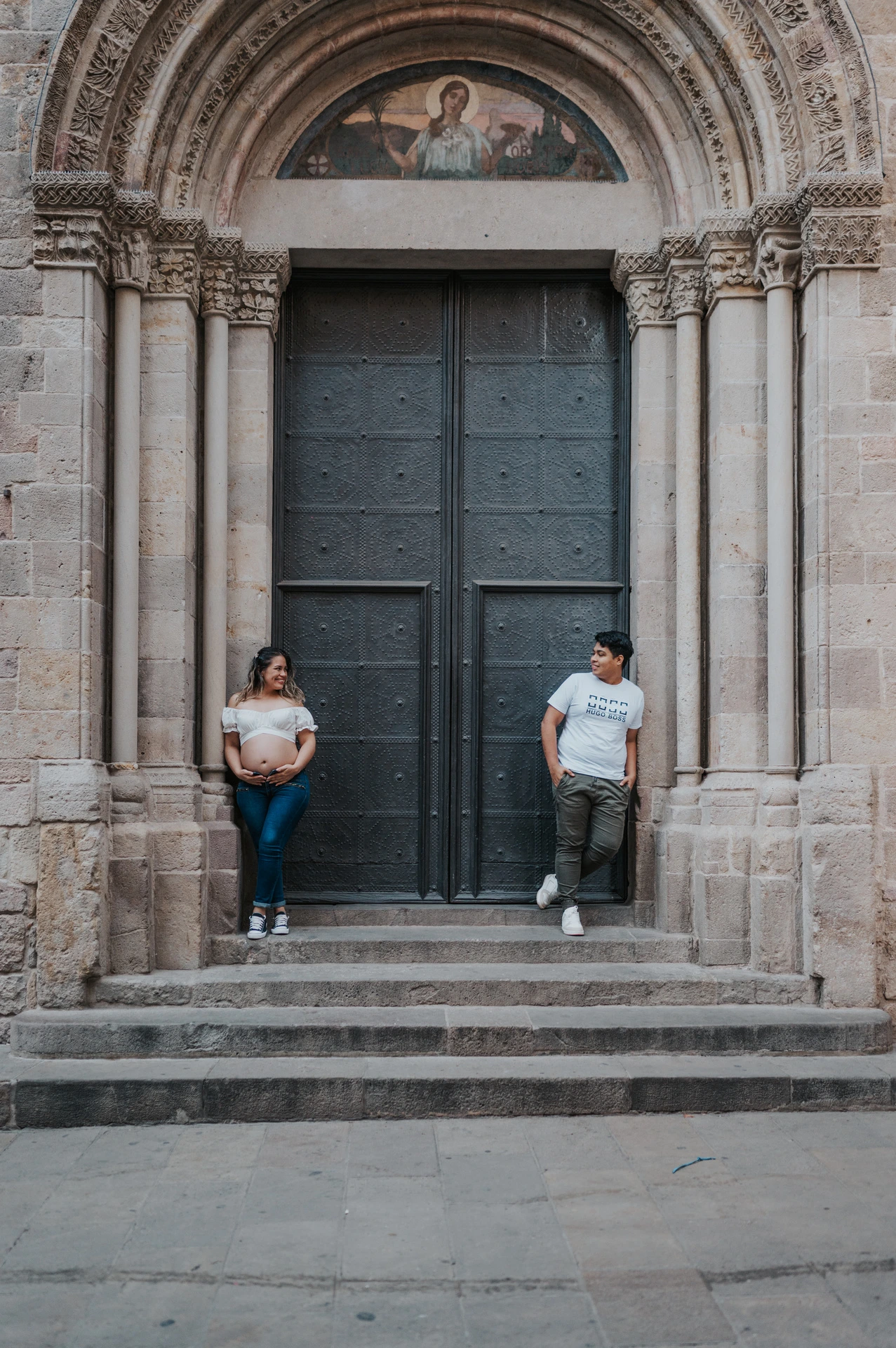 The couple standing on either side of a large doorway framed by carved stone and steps during their maternity photoshoot in Barcelona.