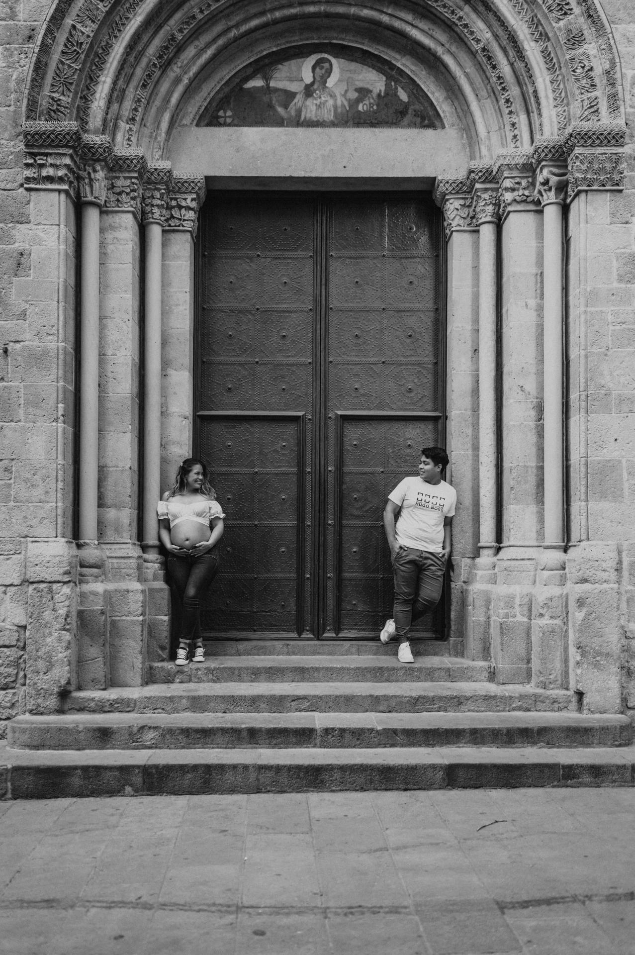 Black-and-white photograph: the couple standing on either side of a large doorway framed by carved stone and steps during their maternity photoshoot in Barcelona.