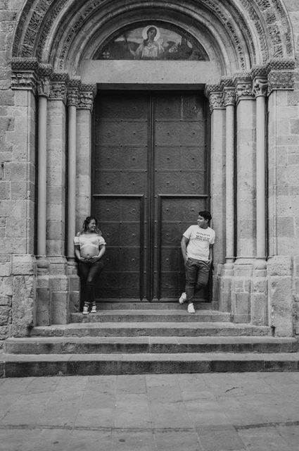 Together beside the carved stone doorway in black and white | Barcelona maternity photoshoot Black-and-white photograph: the couple standing on either side of a large doorway framed by carved stone and steps during their maternity photoshoot in Barcelona.