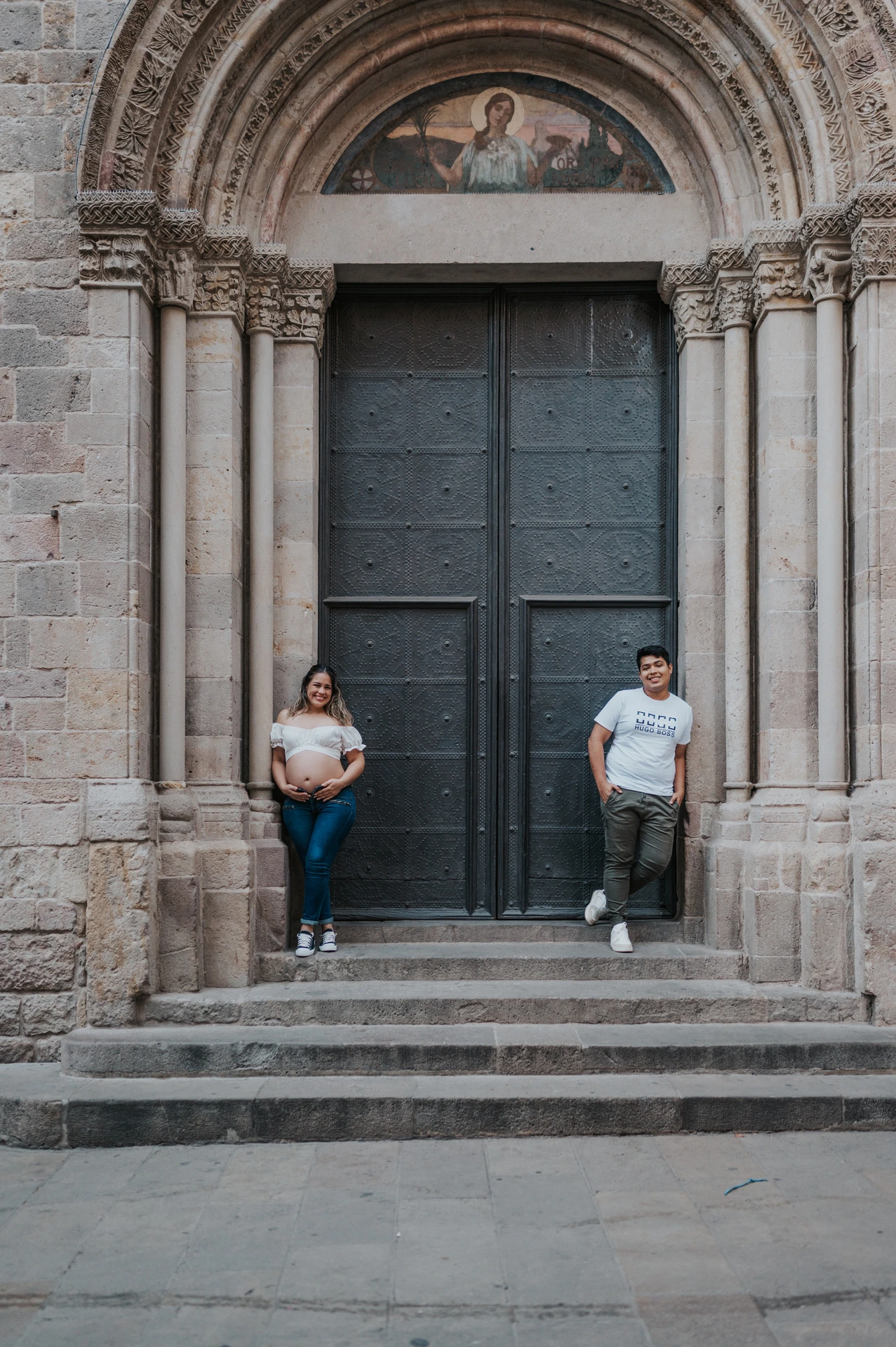 The couple standing on either side of a large doorway framed by carved stone and steps during their maternity photoshoot in Barcelona.