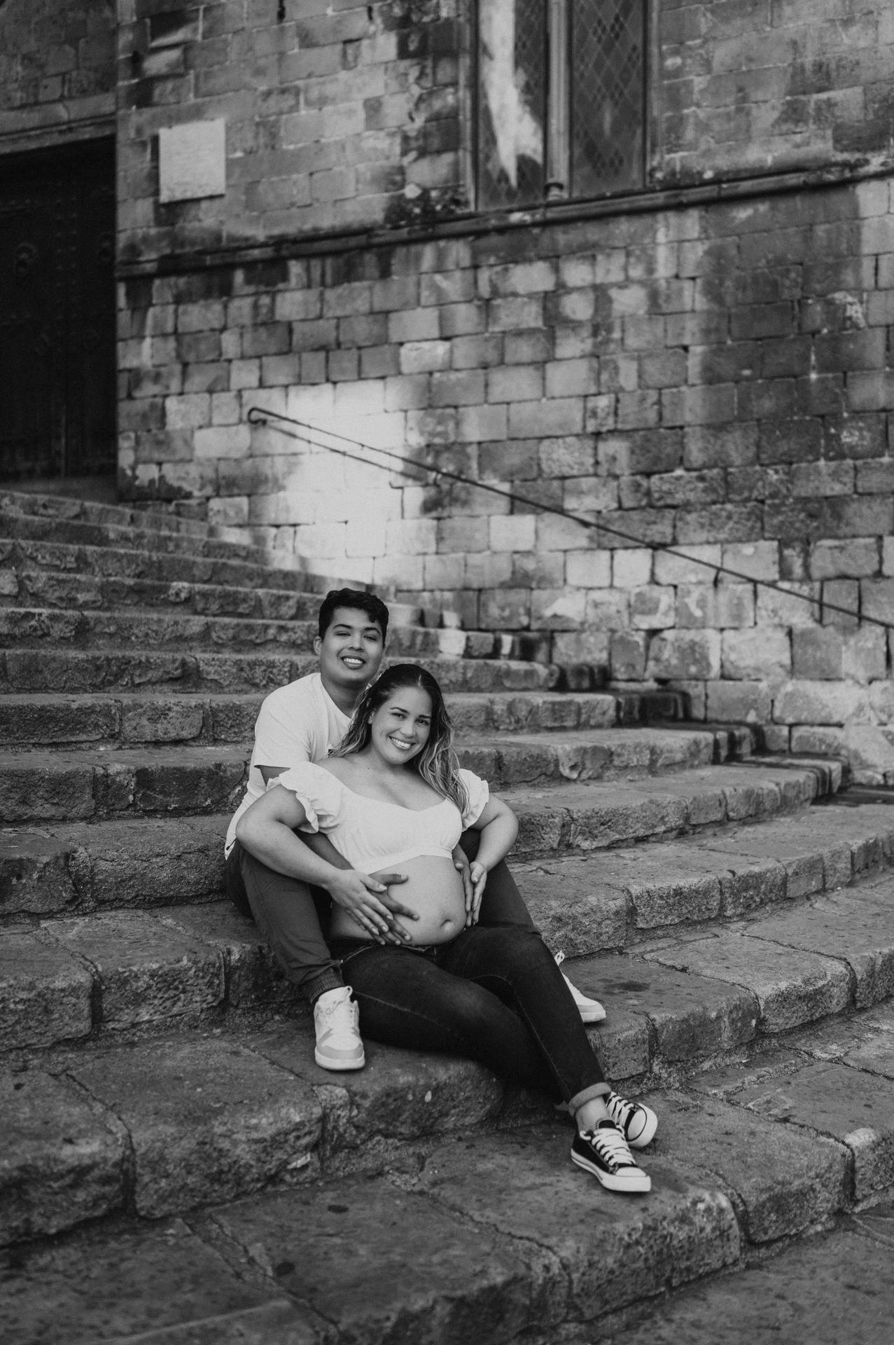 Black-and-white photograph: the couple sitting close together on the stone steps and smiling towards the camera during their maternity photoshoot in Barcelona.