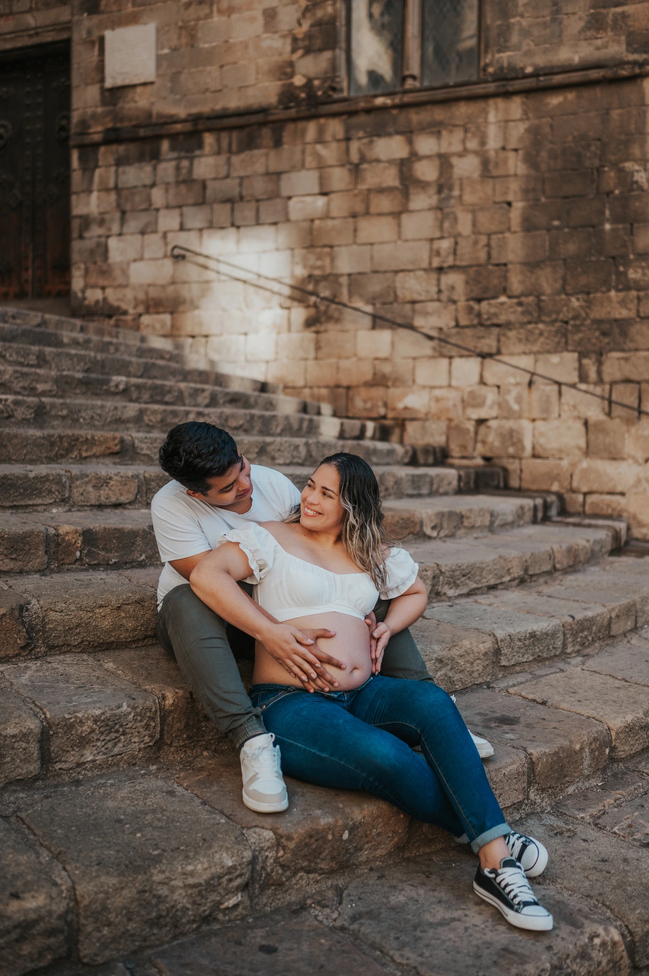 The couple sitting together on the stone steps with their hands resting on her bump during their maternity photoshoot in Barcelona.